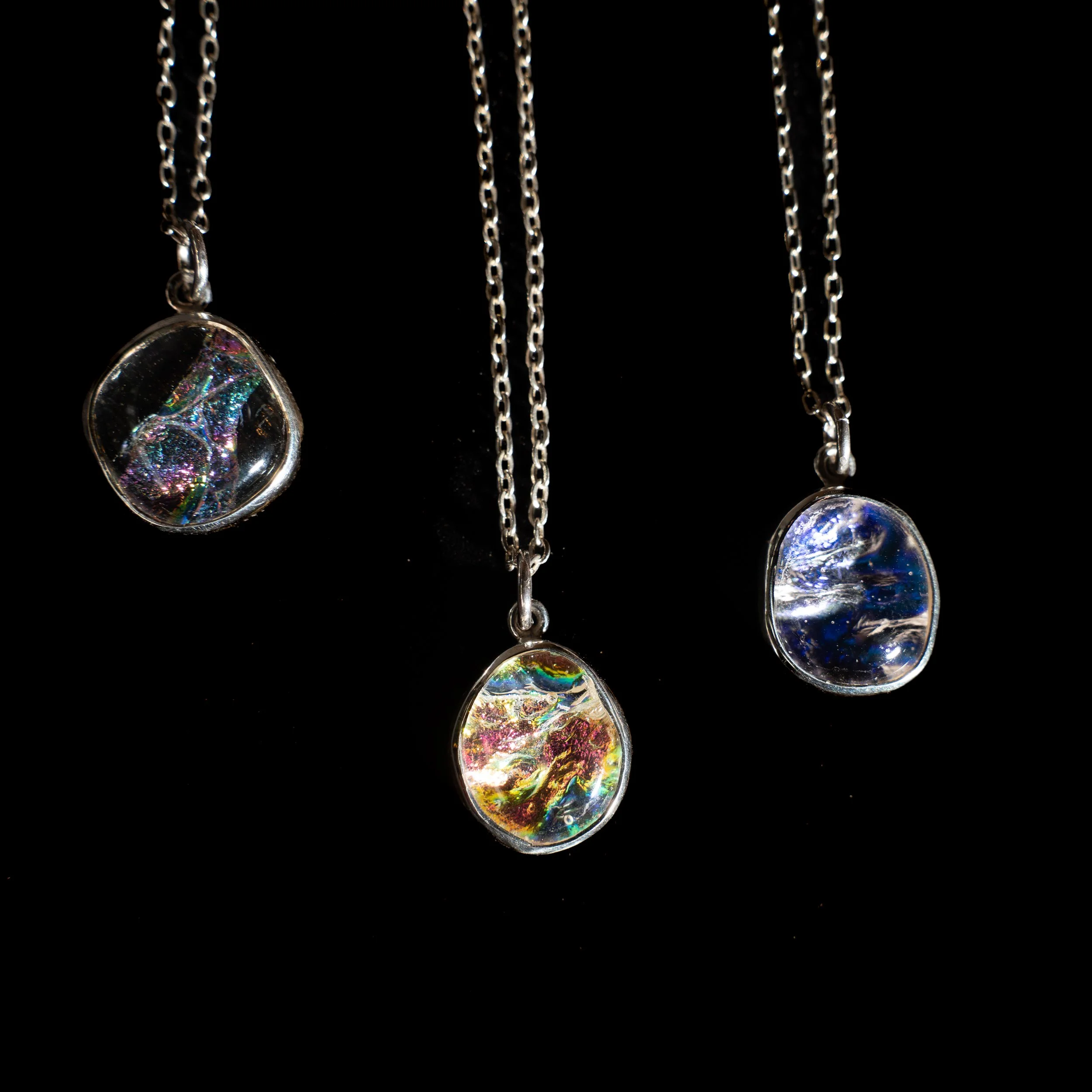 Cuatro collares con colgantes de vidrio con efectos de luz en diferentes colores, colgados en cadenas de metal sobre fondo negro.