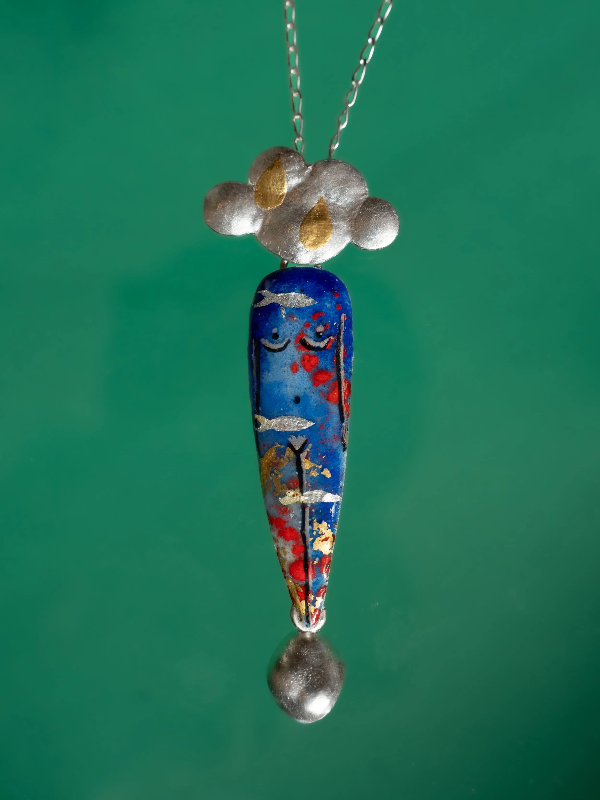 Collar con cadena y colgante en forma de figura antropomorfa con color azul y detalles en dorado, con gotas de agua en la superficie.