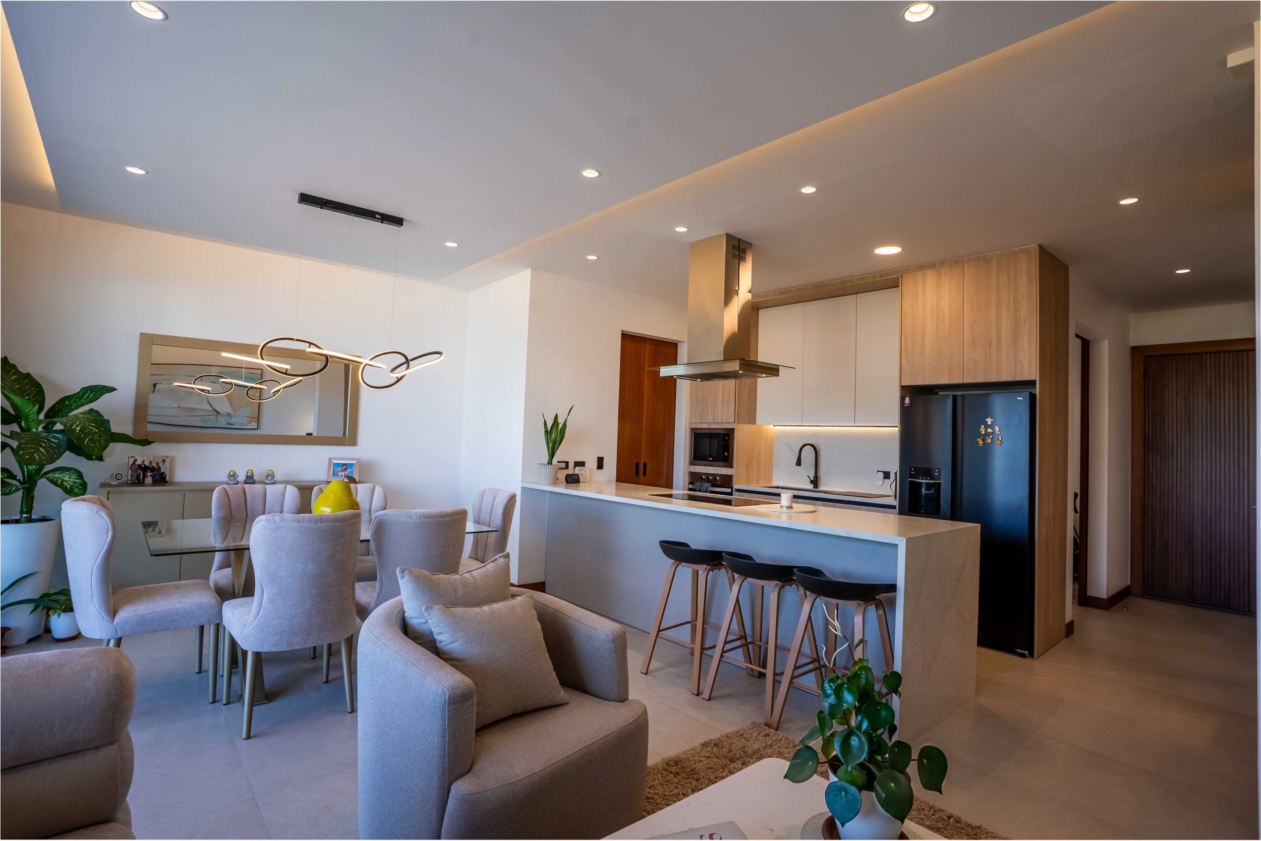 Interiores de una sala y cocina modernas con muebles beige y madera, plantas y una iluminación cálida.