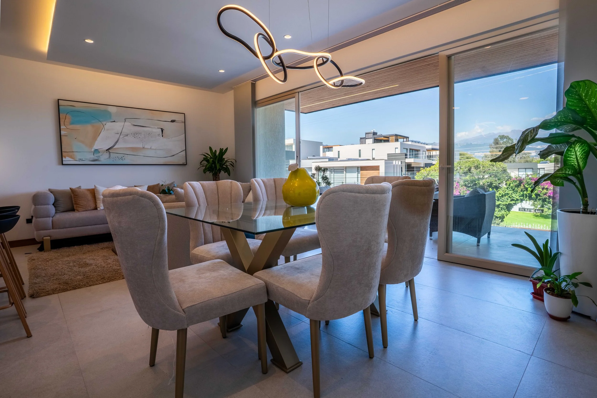 Sala de estar moderna con sillones beige, mesa de comedor de vidrio, un jarrón amarillo y plantas verdes, con ventanas grandes que ofrecen vista a edificios y un cielo azul.