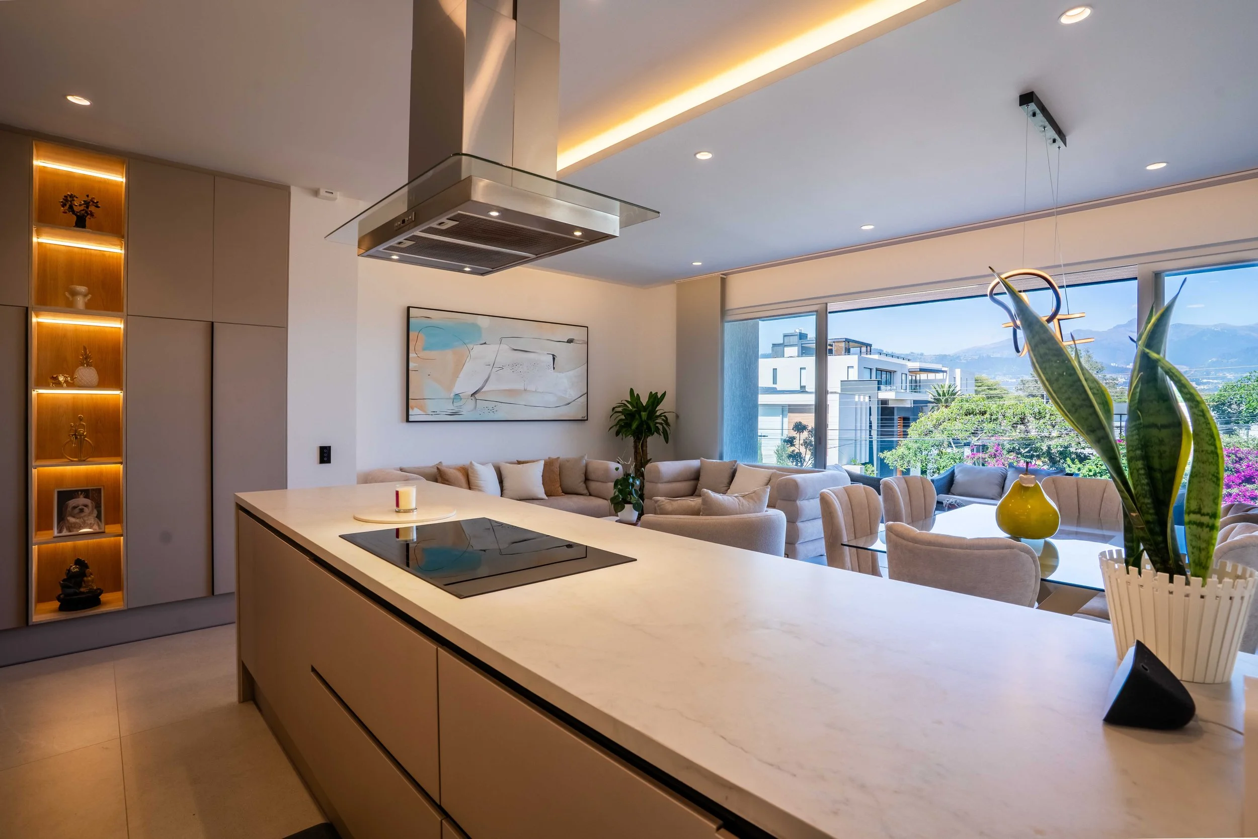 Interiores de una sala moderna con ventanas grandes, vista a edificios y montañas, decoración minimalista, plantas y una pintura abstracta en la pared.