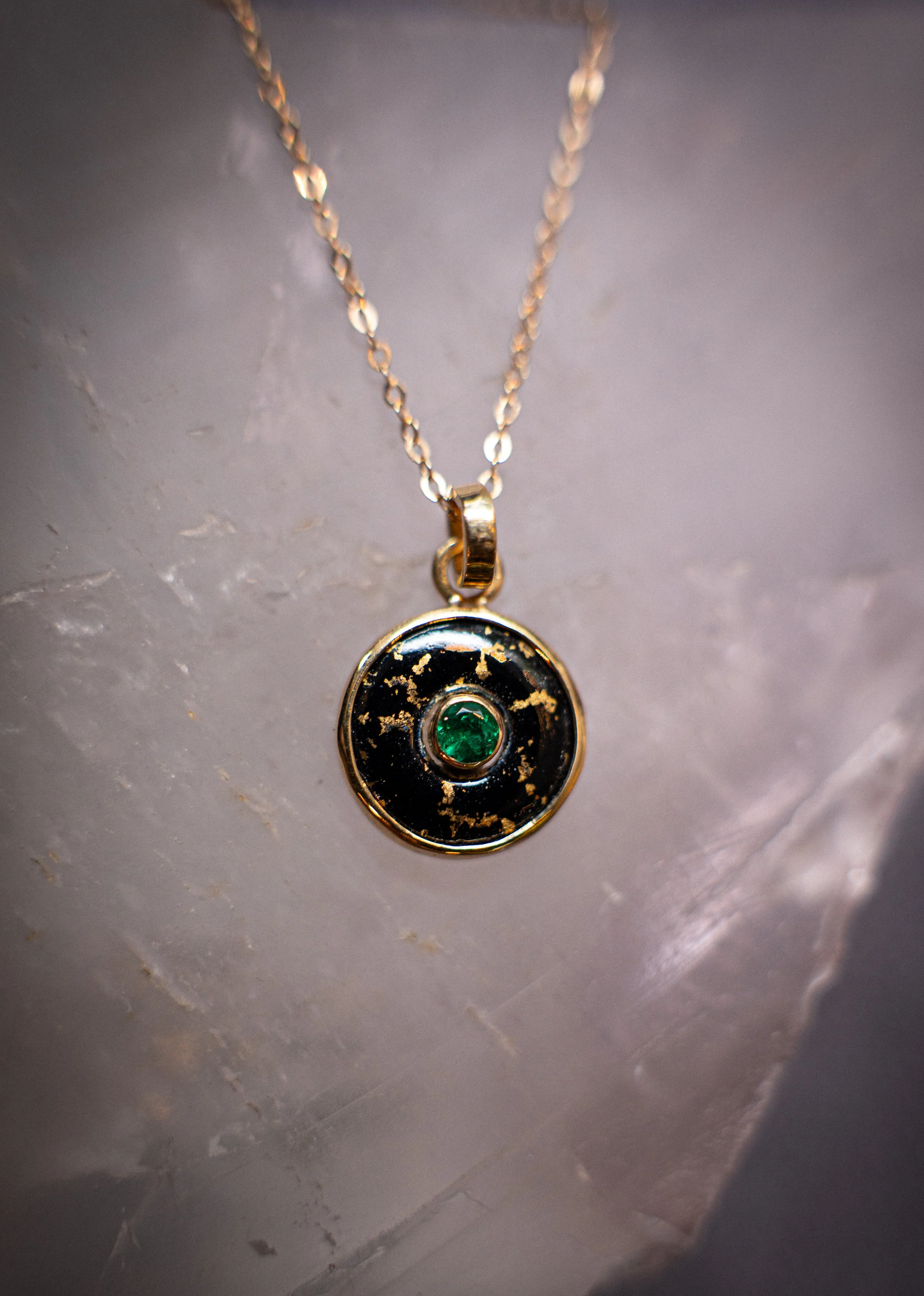 Un collar con un colgante redondo, dorado con detalles en negro y una piedra verde en el centro, colgado de una cadena dorada.