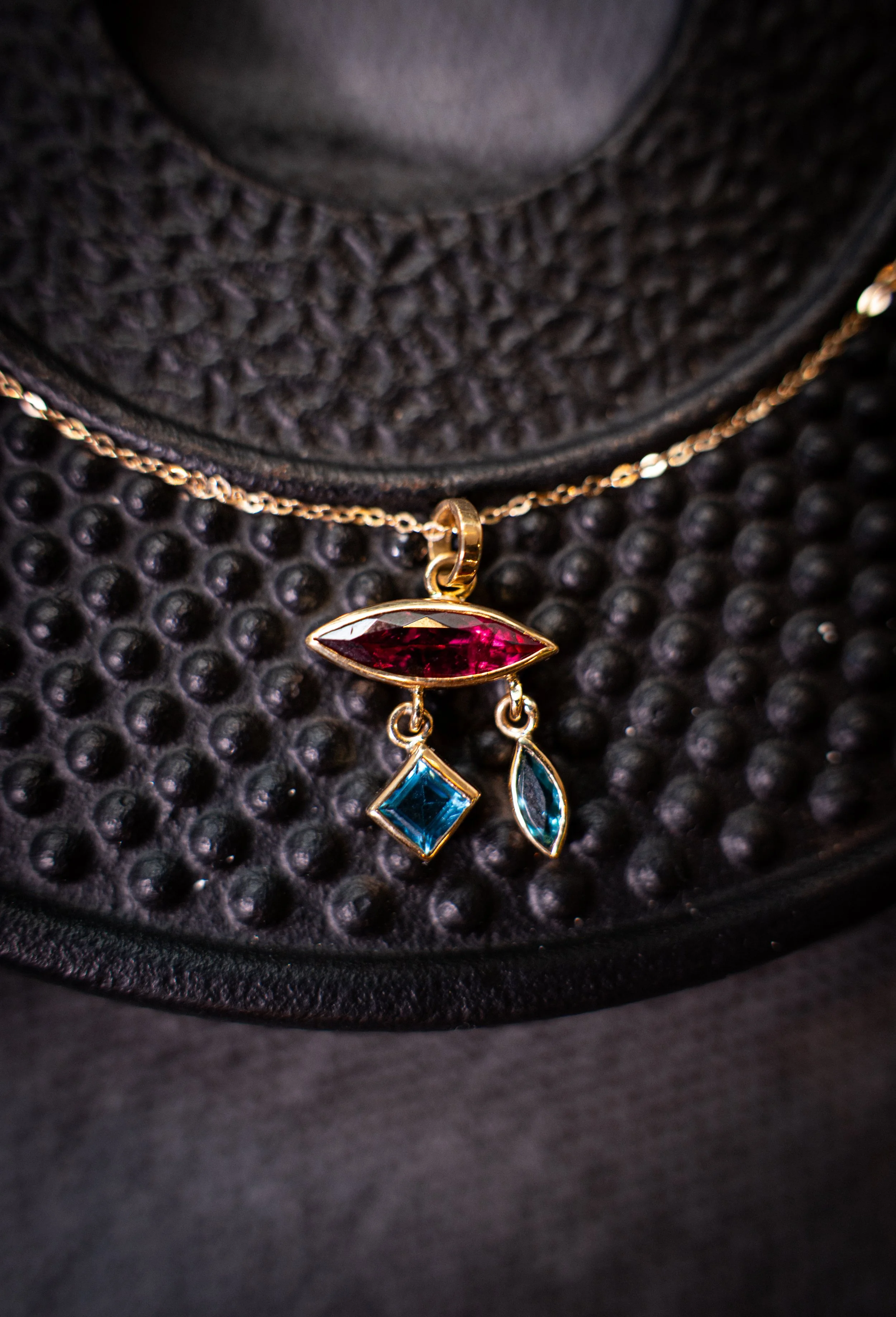 Cadena con colgante con piedras de colores, suspendida frente a una superficie de cuero negra con textura de burbujas.