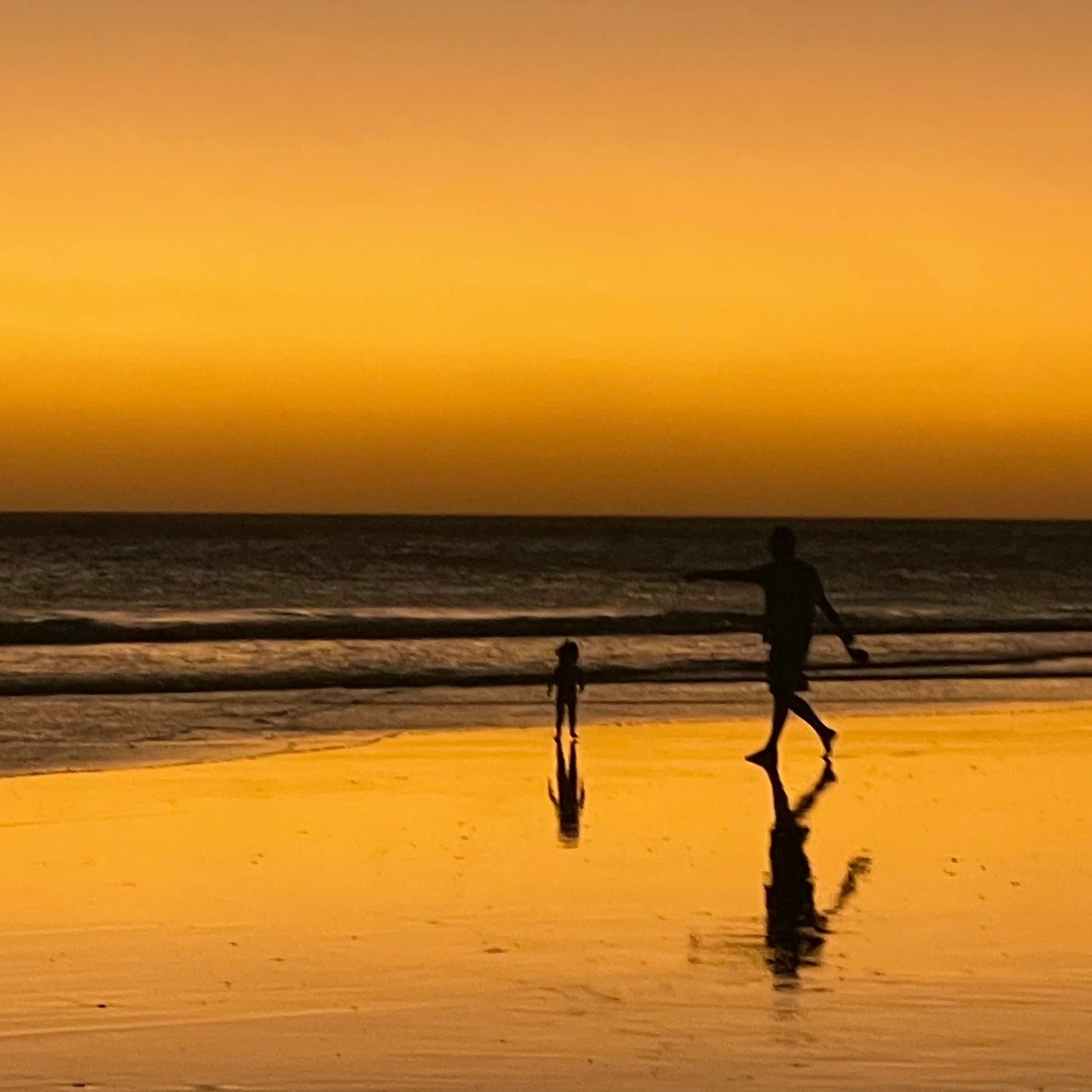 A beautiful orange sunset over the ocean with a beach in the foreground. There are two people walking on the beach.