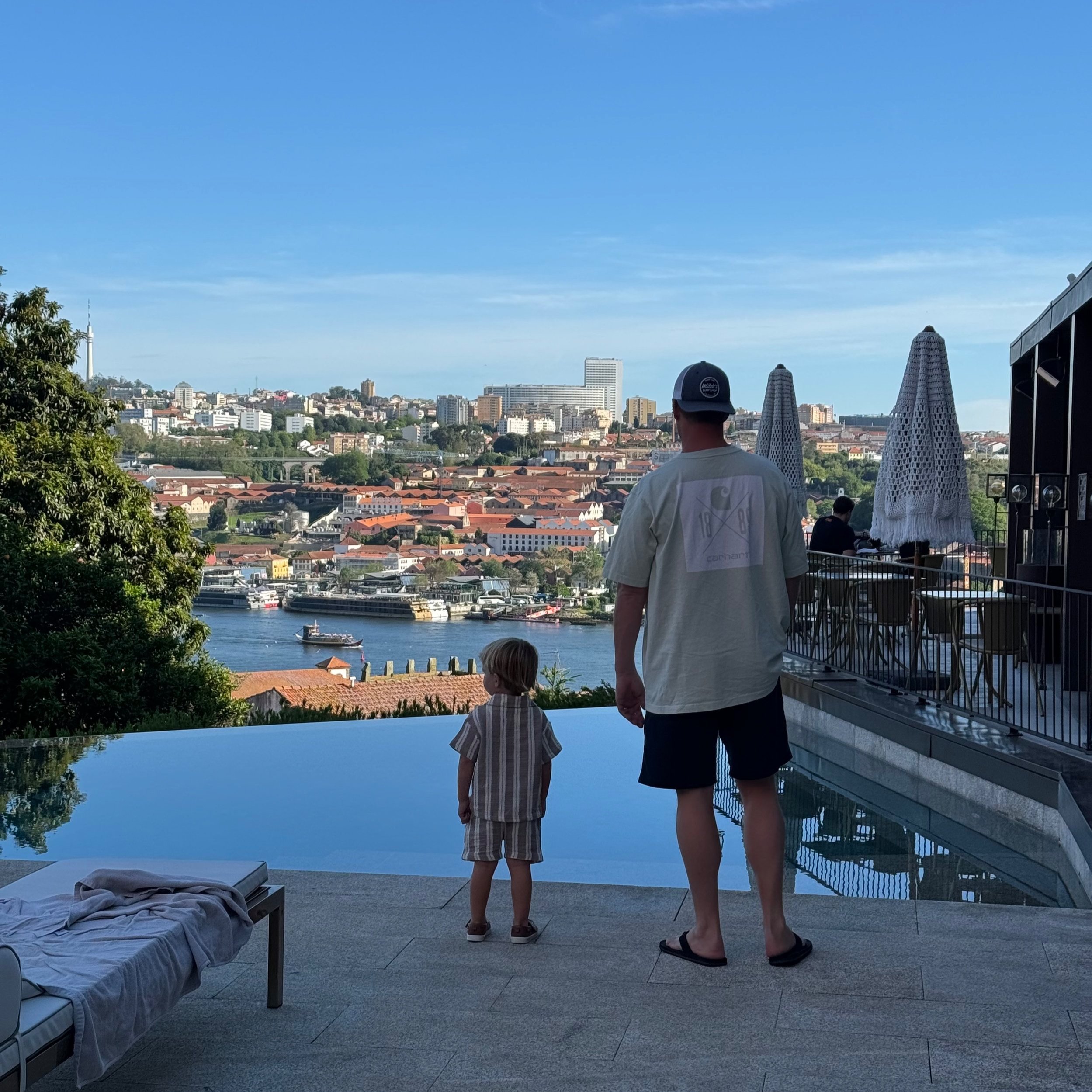 A rooftop pool area overlooking a city skyline with a river, boats, and high-rise buildings on a clear day. Two people, a man and a child, stand near the pool, looking at the view, with folded umbrellas and a bed with a towel in the scene.