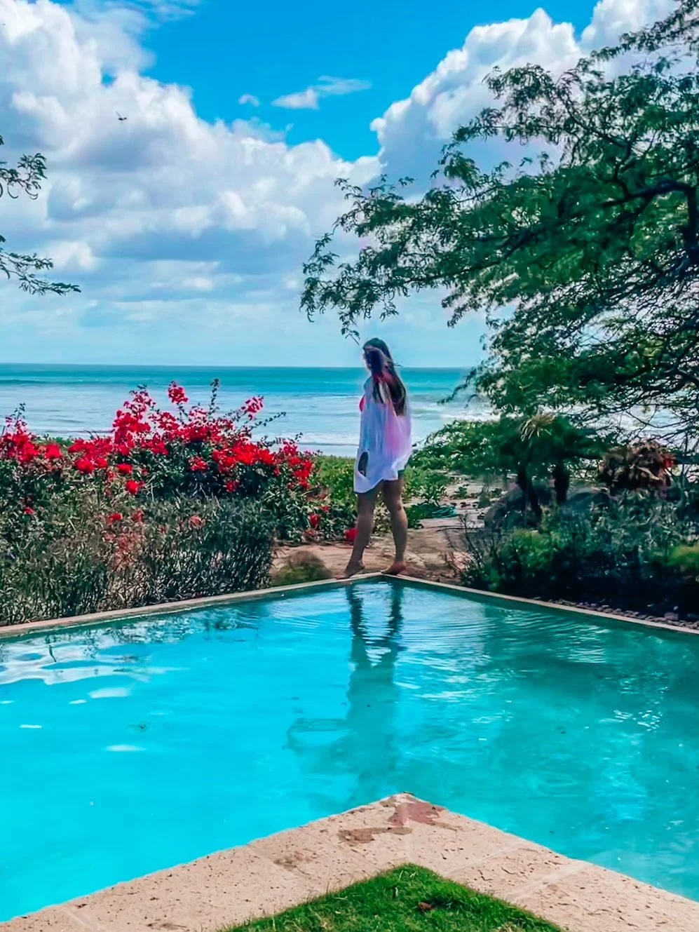 A woman standing by a swimming pool with an ocean view, surrounded by green trees and pink flowers, under a partly cloudy sky.