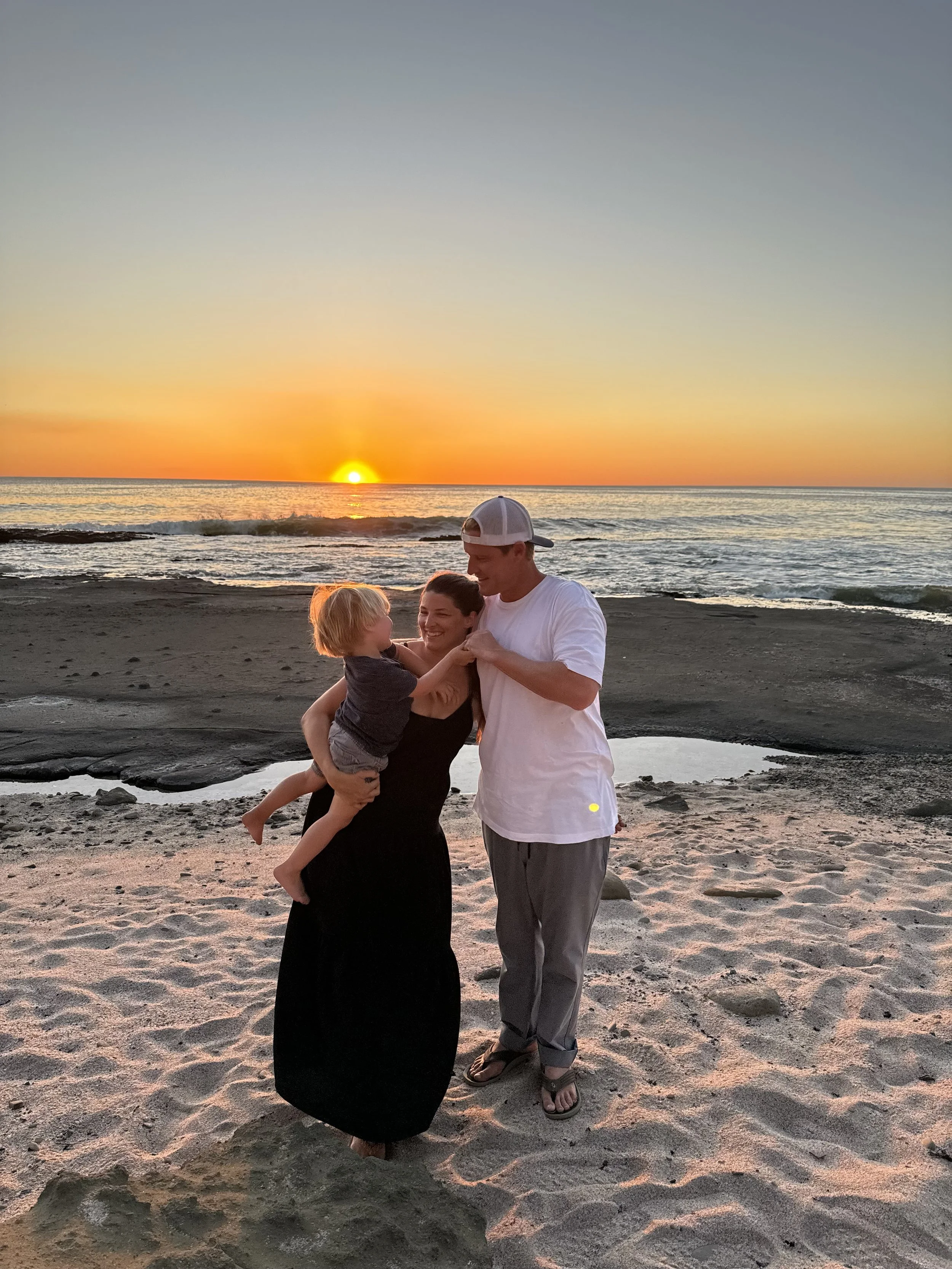 A family of three, including a mother, father, and young boy, standing on a beach at sunset with waves in the background. The mother is holding the boy, and the father is smiling, all enjoying each other's company.