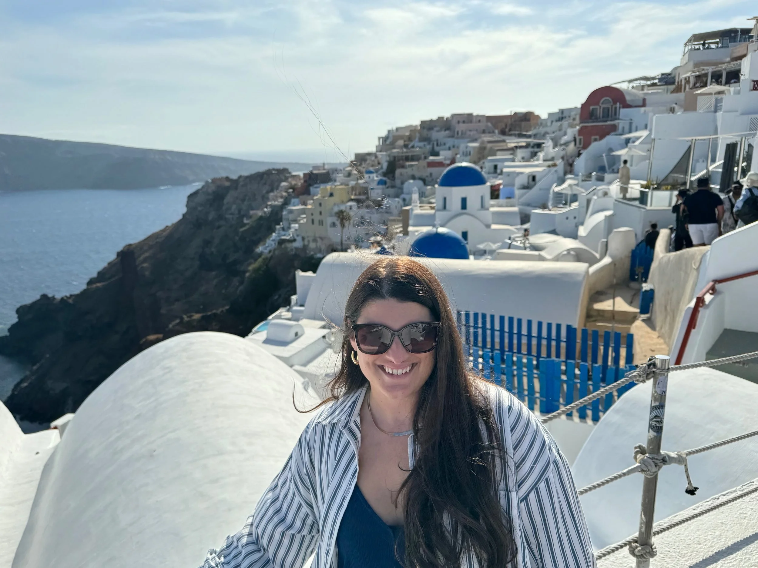 Smiling woman with long dark hair wearing sunglasses and a striped shirt at a scenic Mediterranean coastal village with white buildings and blue-domed churches.