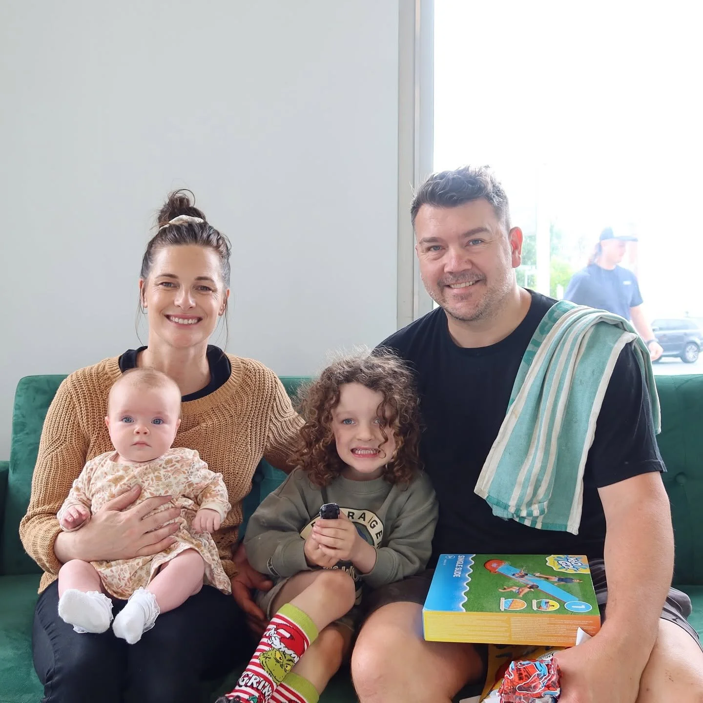 A family of four sitting on a green couch at a restaurant, smiling at the camera. The woman holds a baby with light skin, and a child with curly hair and light skin is seated between the man and the woman. The man has a towel over his shoulder, and a board game box is on his lap.