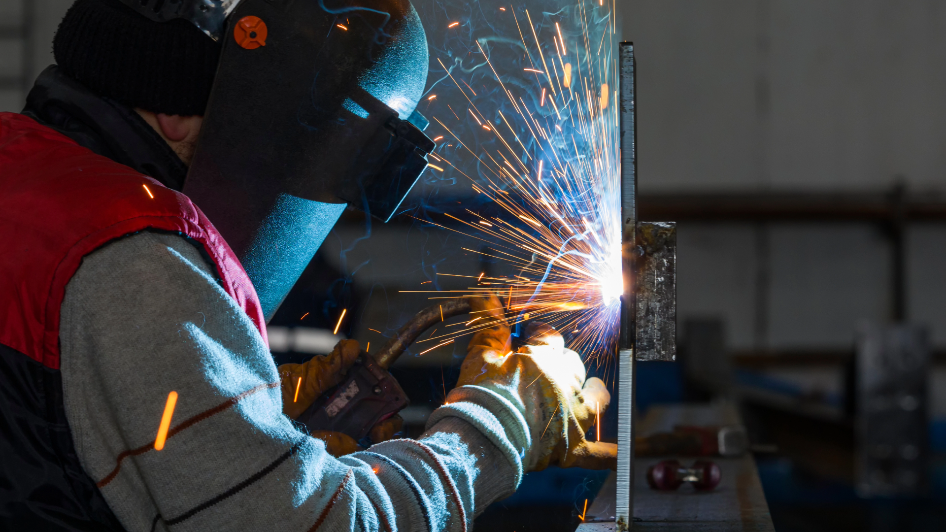 A person welding metal in an industrial workshop, wearing protective gear including a welding helmet and gloves, with sparks flying from the welding process.