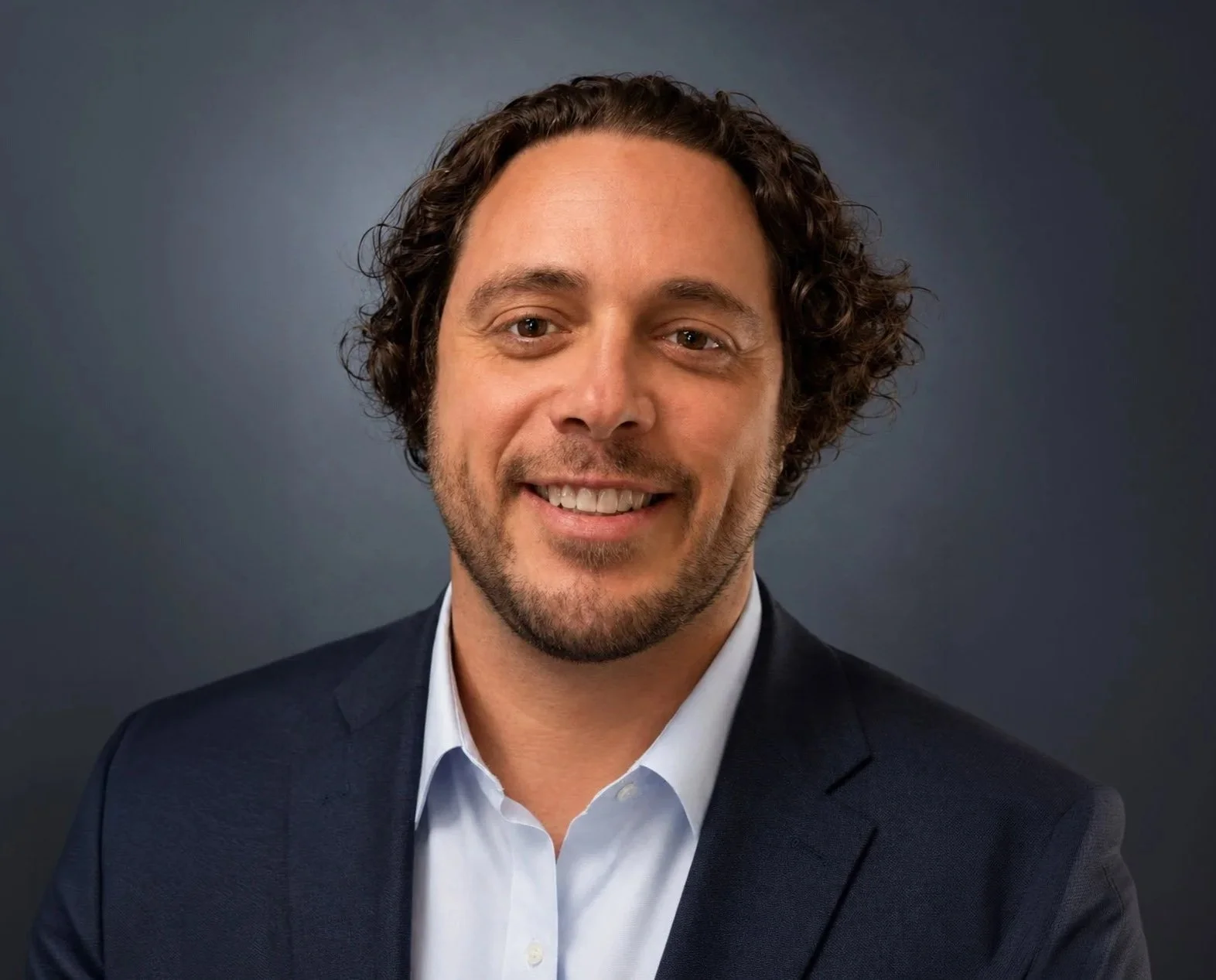 A smiling man with curly brown hair, wearing a dark suit jacket and a white shirt, against a dark background.