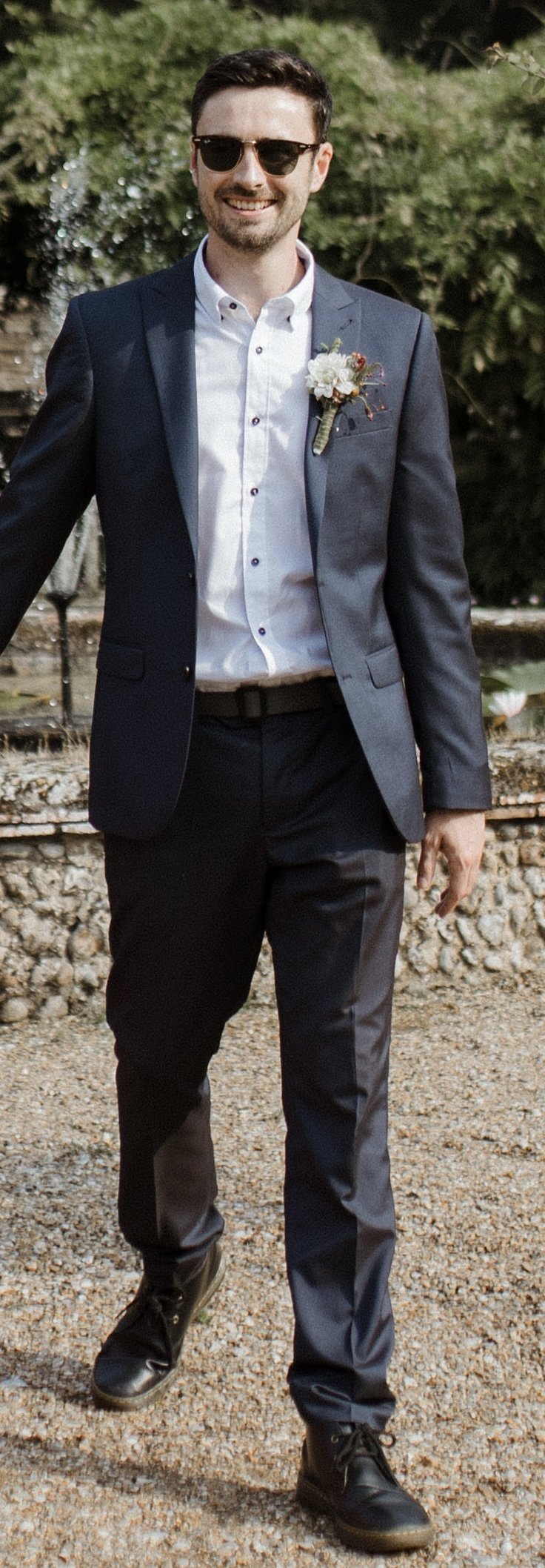 A smiling man wearing sunglasses, a dark suit with a boutonniere, white shirt, and black shoes standing outdoors on a gravel path.