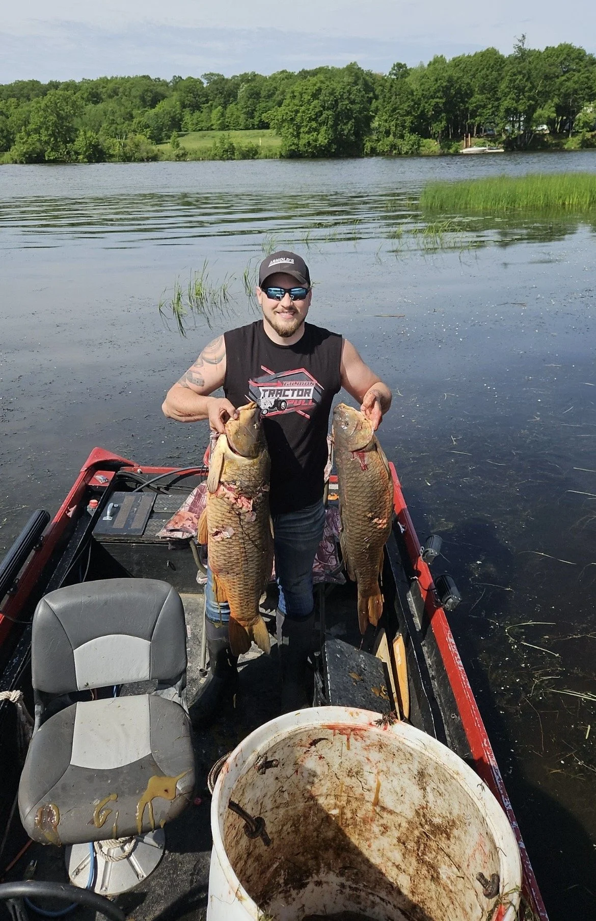 A man on a boat holding two large fish, with a lake and trees in the background.