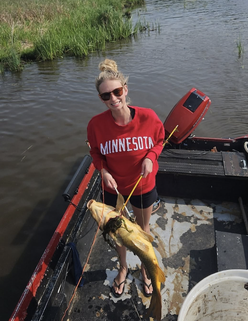 A woman standing on a boat holding a large fish she caught, smiling, wearing sunglasses and a red Minnesota sweatshirt, with water and grass in the background.