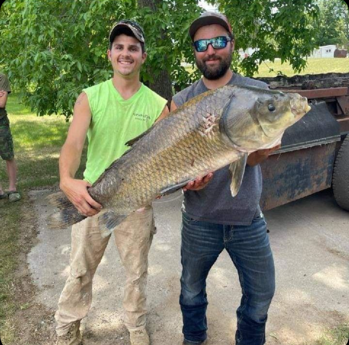 Two men outdoors holding a large fish with green trees and a trailer in the background.