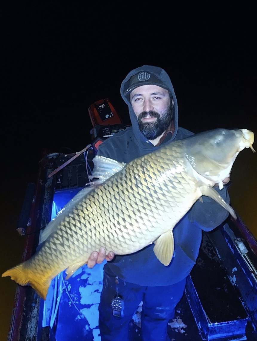 Man holding a large fish on a boat at night with a dark sky.