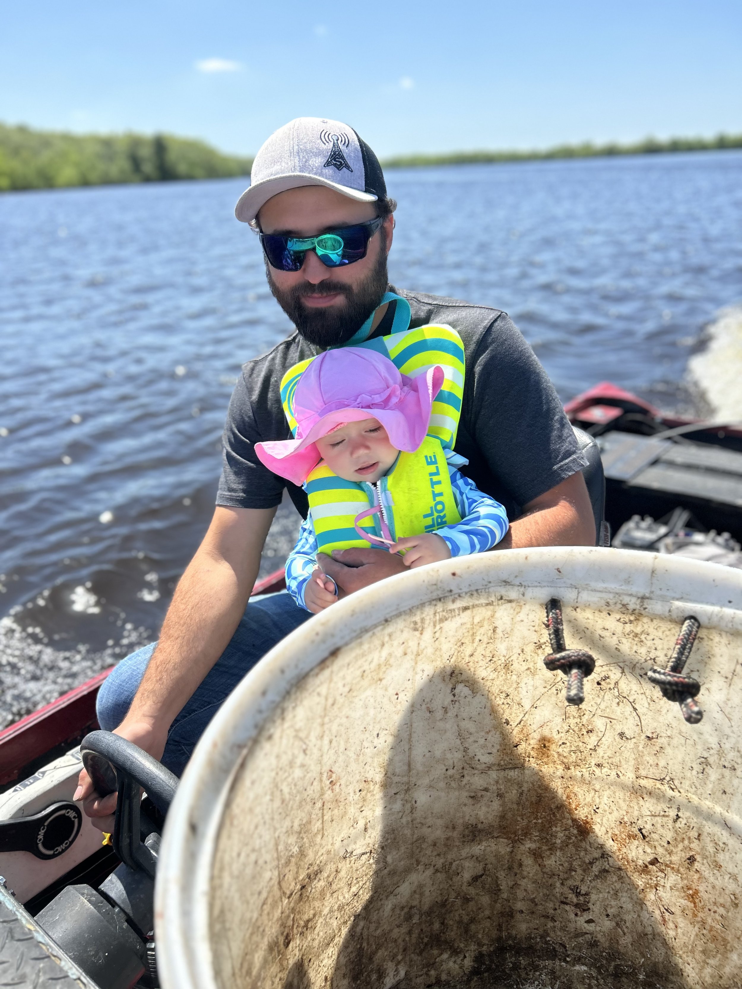 A man wearing sunglasses and a white cap holding a young child with a pink hat and a life jacket, sitting on a boat with water and trees in the background.