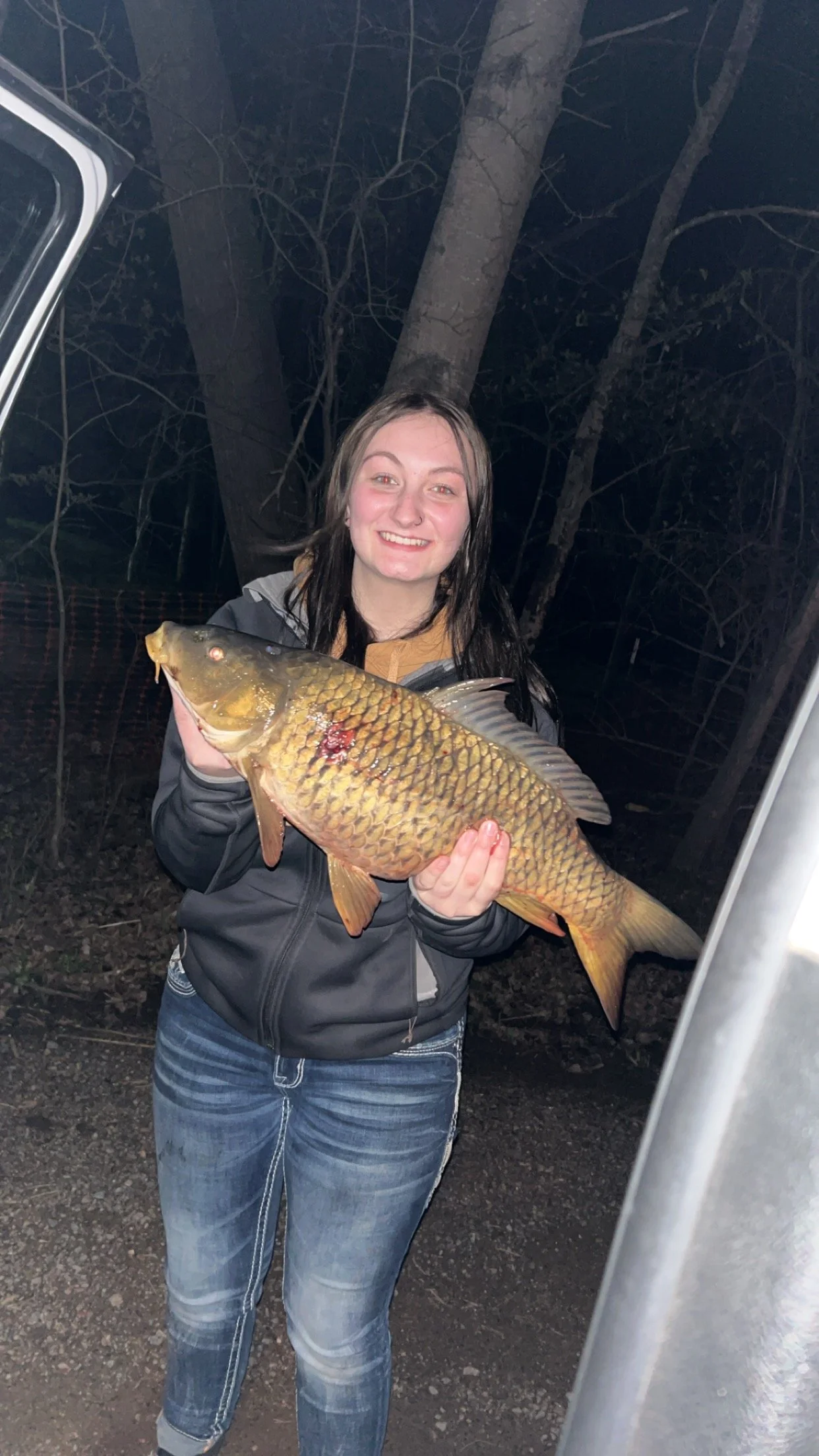 A woman holding a large fish in an outdoor setting at night, with trees in the background.