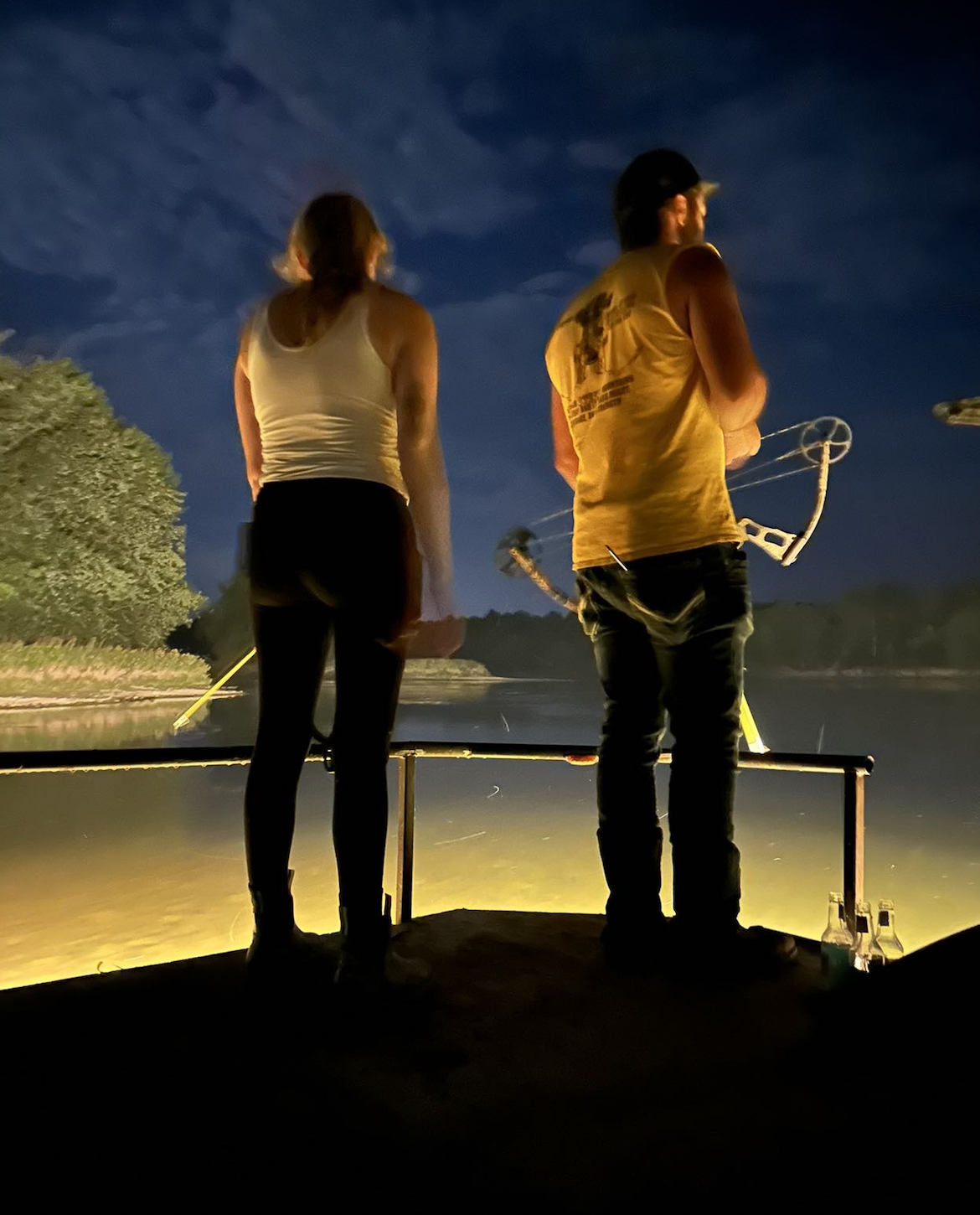 Two people stand on a boat at night, fishing in a river with a dark sky and clouds in the background.