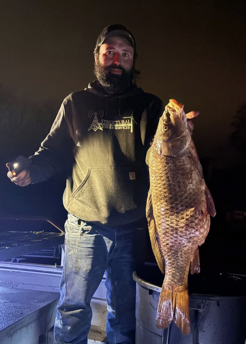 A man with a beard and mustache, dressed in a dark hoodie with a logo and jeans, holding a large fish at night on a boat.