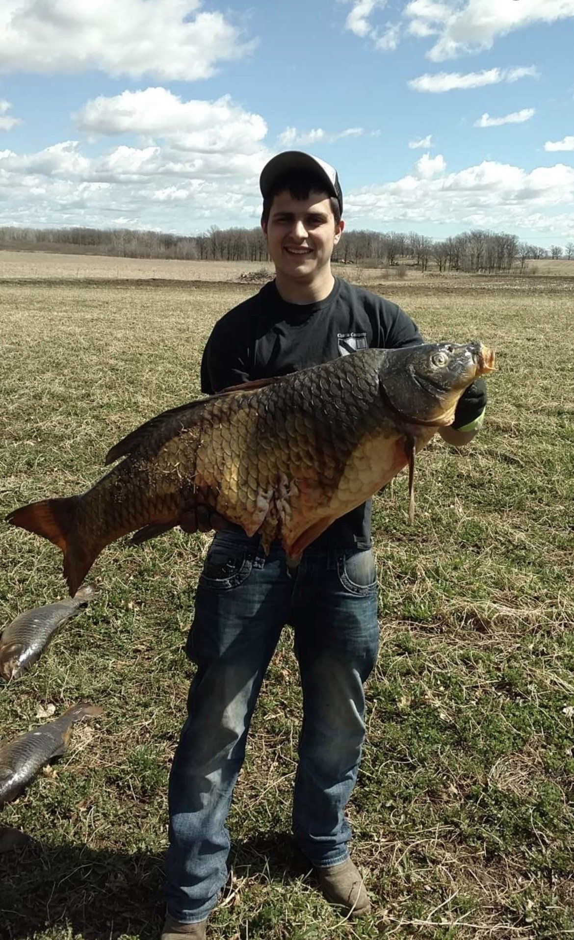 A young man standing in an open field holding a large fish. He is wearing a black t-shirt, jeans, and a cap, smiling at the camera. The sky is partly cloudy.