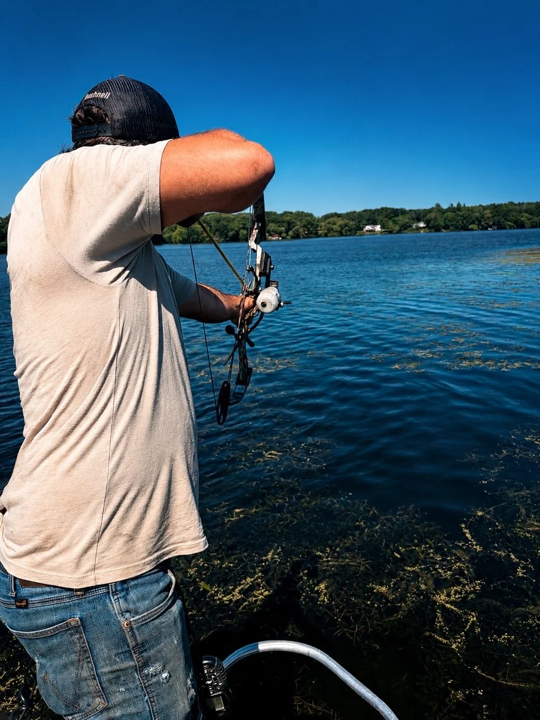 Man fishing by a lake on a sunny day, holding a bow with a fishing line, wearing a black cap and casual clothing.