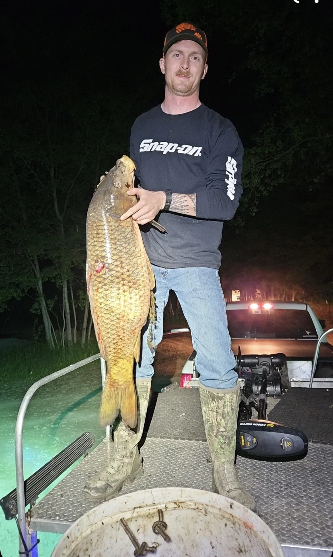 Man standing on boat holding large fish he caught at night, with fishing gear and vehicles in the background.