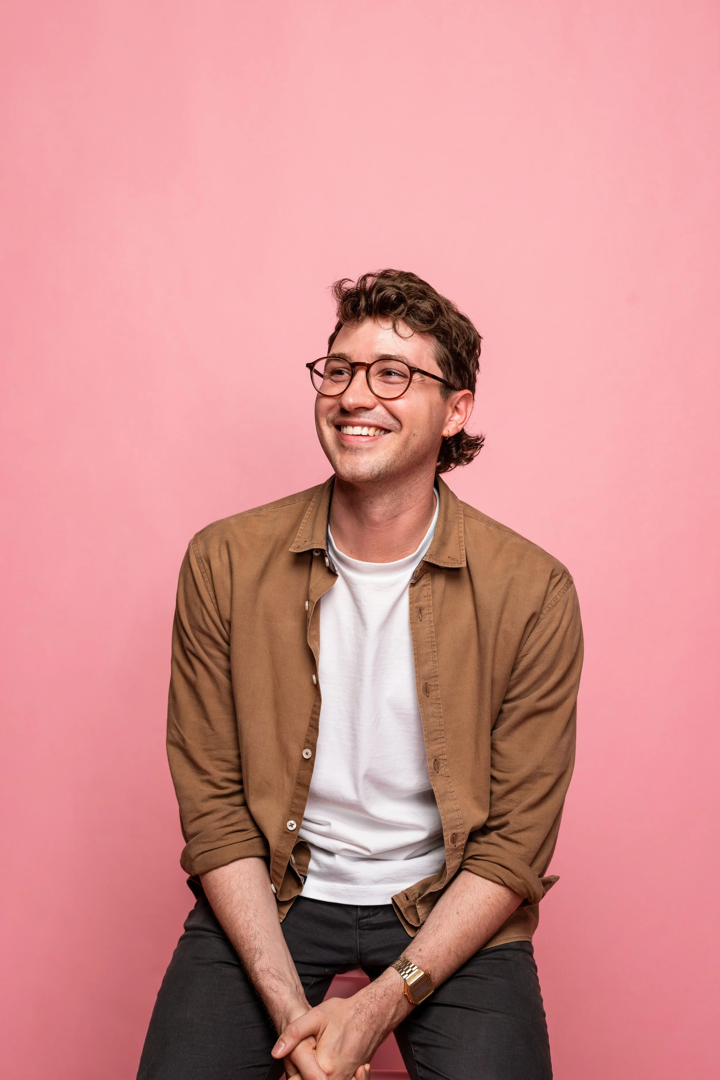 A young man with glasses, curly brown hair, and a light complexion, smiling and looking to the side, sitting against a pink background. He is wearing a white t-shirt, a tan button-up shirt, and a gold wristwatch.