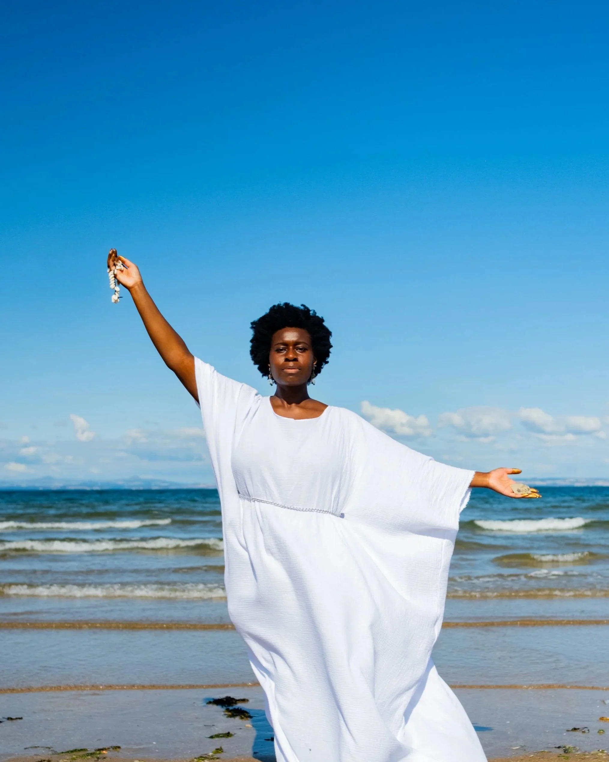 Garen, a Black woman in her thirties, faces us with her right arm upraised and her left arm a little extended. Her expression is serene. She is wearing a white robe with a silver chain belt. She has a black Afro.