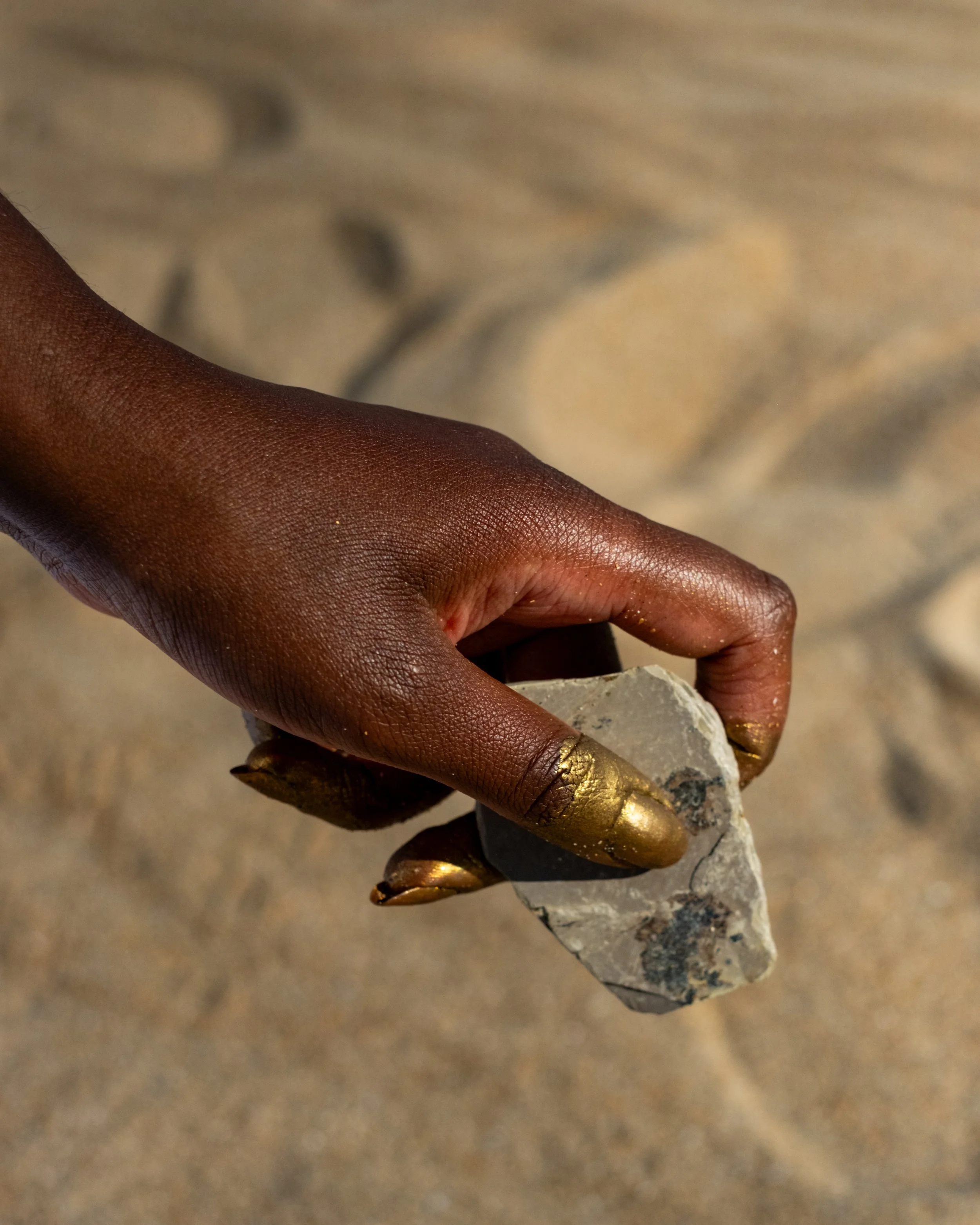 a close up of Garen's hand holding a piece of grey chalk. Her fingertips are painted gold and she is dark-skinned