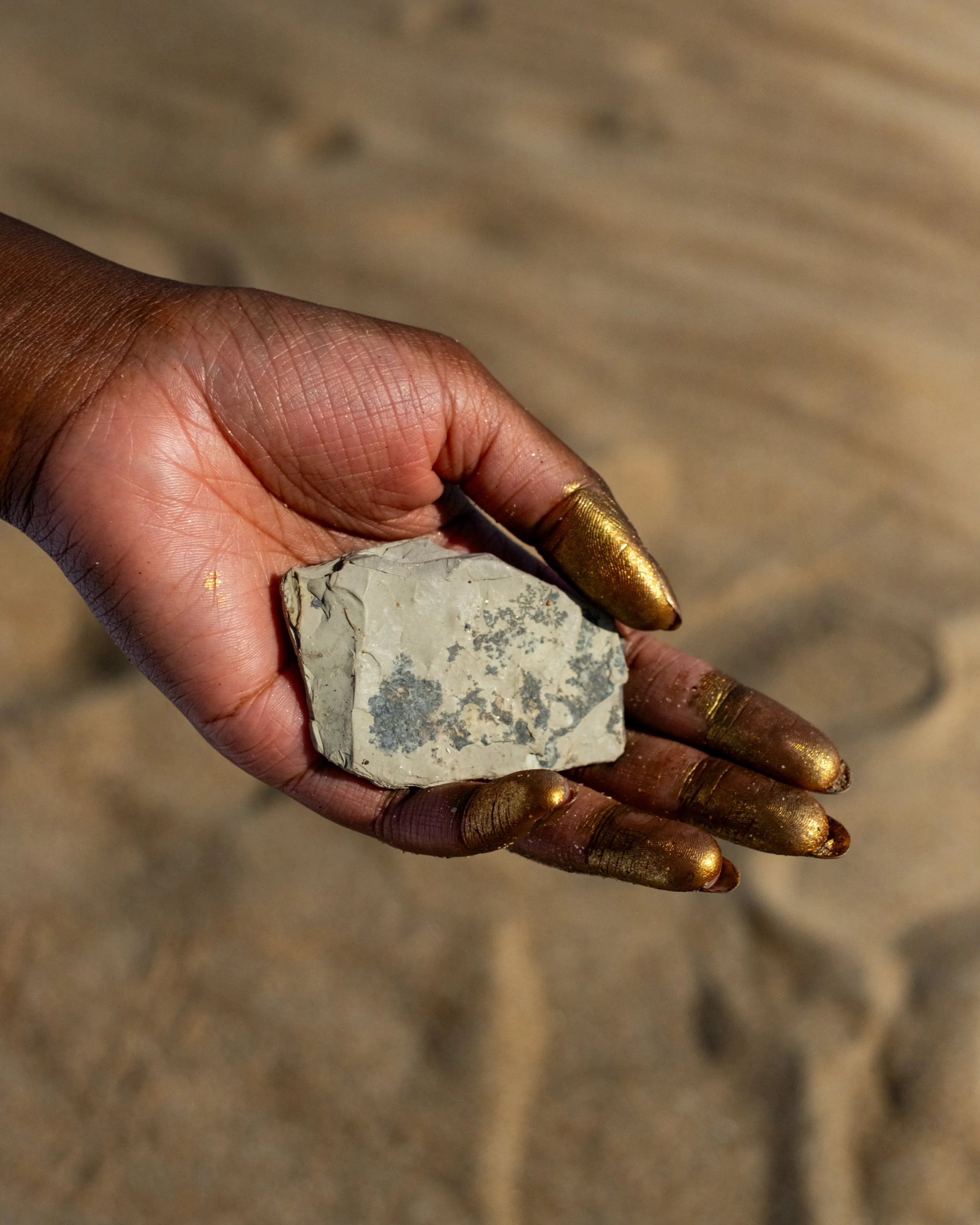 image of Garen's hand holding out a block of grey chalk. Her fingers are tipped with gold paint