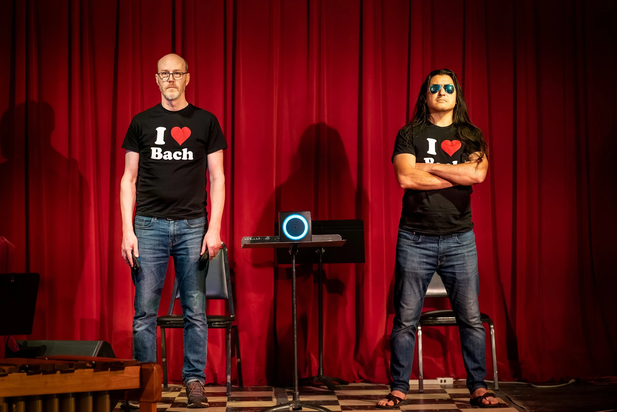 Two men standing on stage in front of a red curtain, wearing black T-shirts that say 'I ❤️ Bach'. The man on the left has glasses, a beard, and light skin, while the man on the right has long dark hair, sunglasses, and lighter skin. There is a music stand and electronic device in front of them.