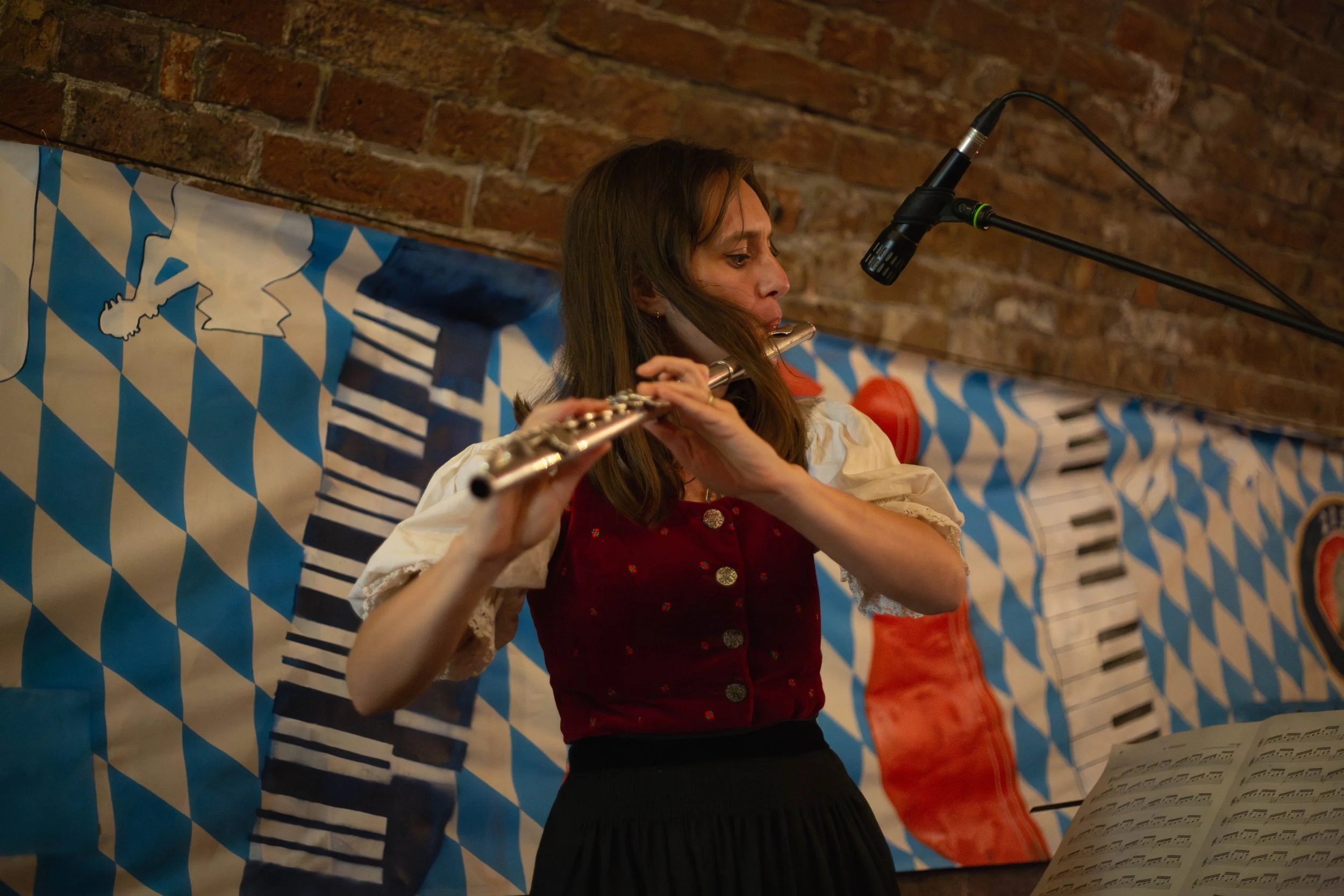 A woman playing a flute in front of a backdrop with Bavarian flag patterns and a piano keyboard design, with a microphone positioned above her.