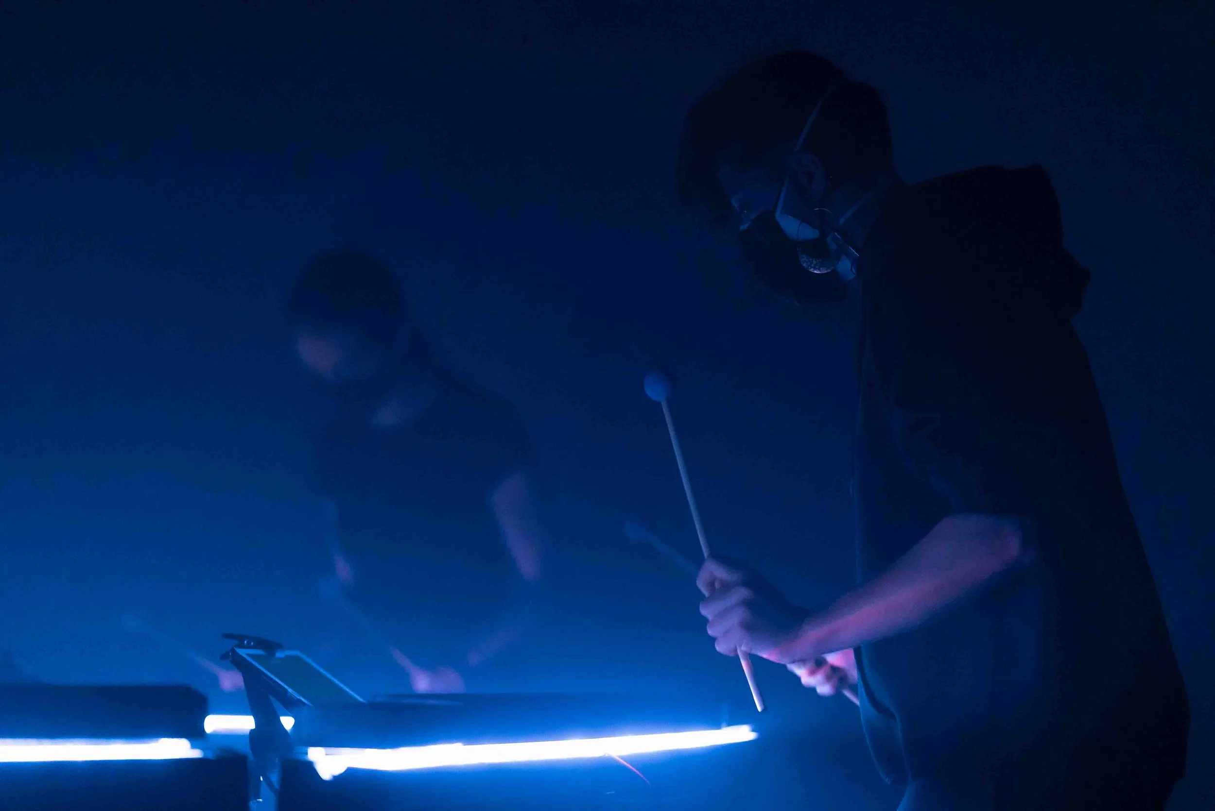 A person wearing glasses and a face mask is playing the piano in a dimly lit room illuminated by blue light.