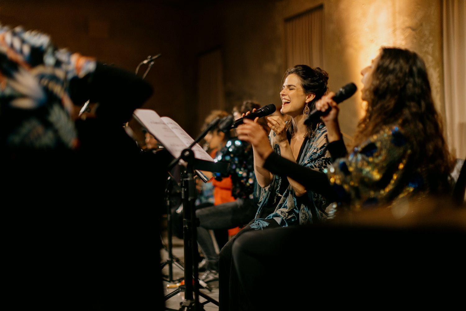 Women singing in a musical performance, holding microphones, with sheet music and a group of musicians in the background.