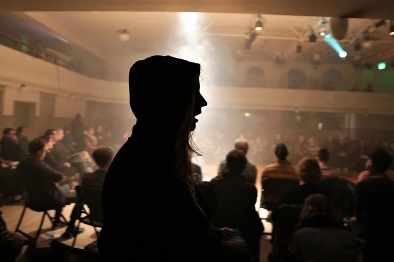 Silhouette of a woman wearing a hooded jacket in front of a large audience seated in an auditorium or theater with stage lighting.