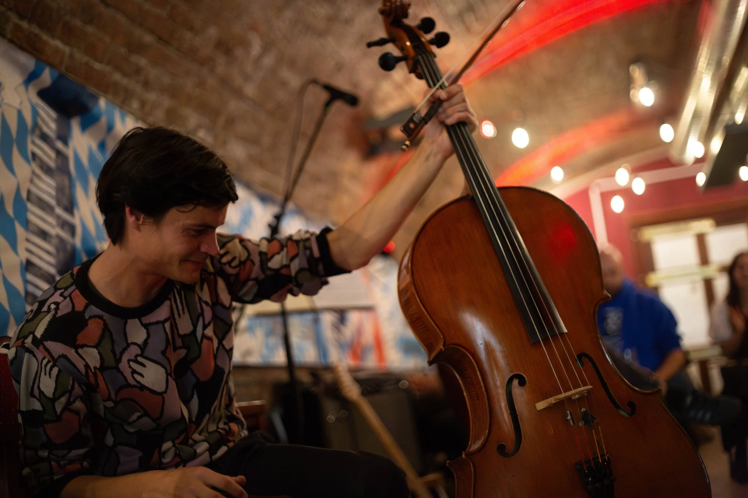 A person with short dark hair playing a cello in an indoor setting with warm lighting and other people in the background.