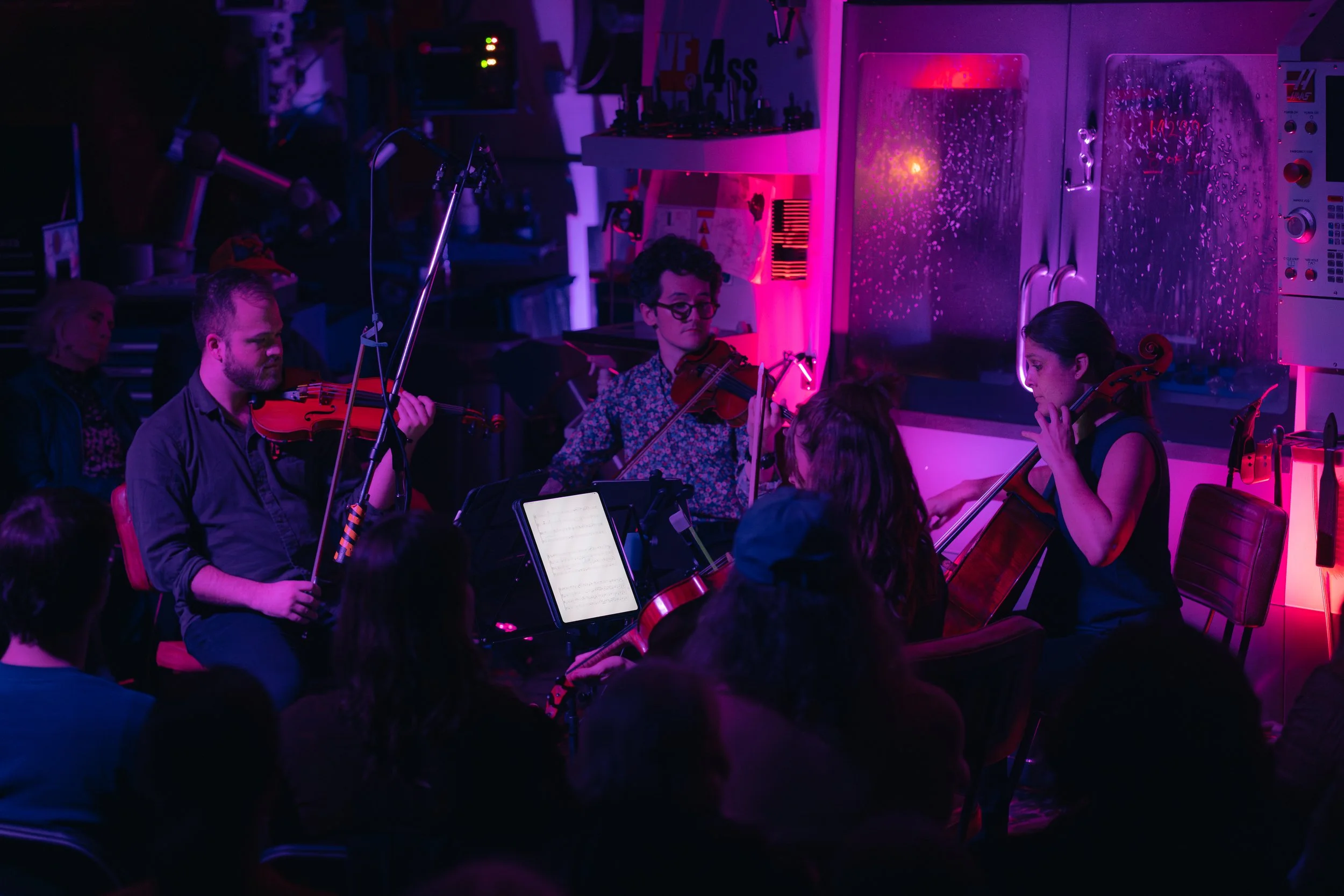 A group of musicians playing violins during a concert illuminated by colorful purple and pink stage lights, with an audience watching.