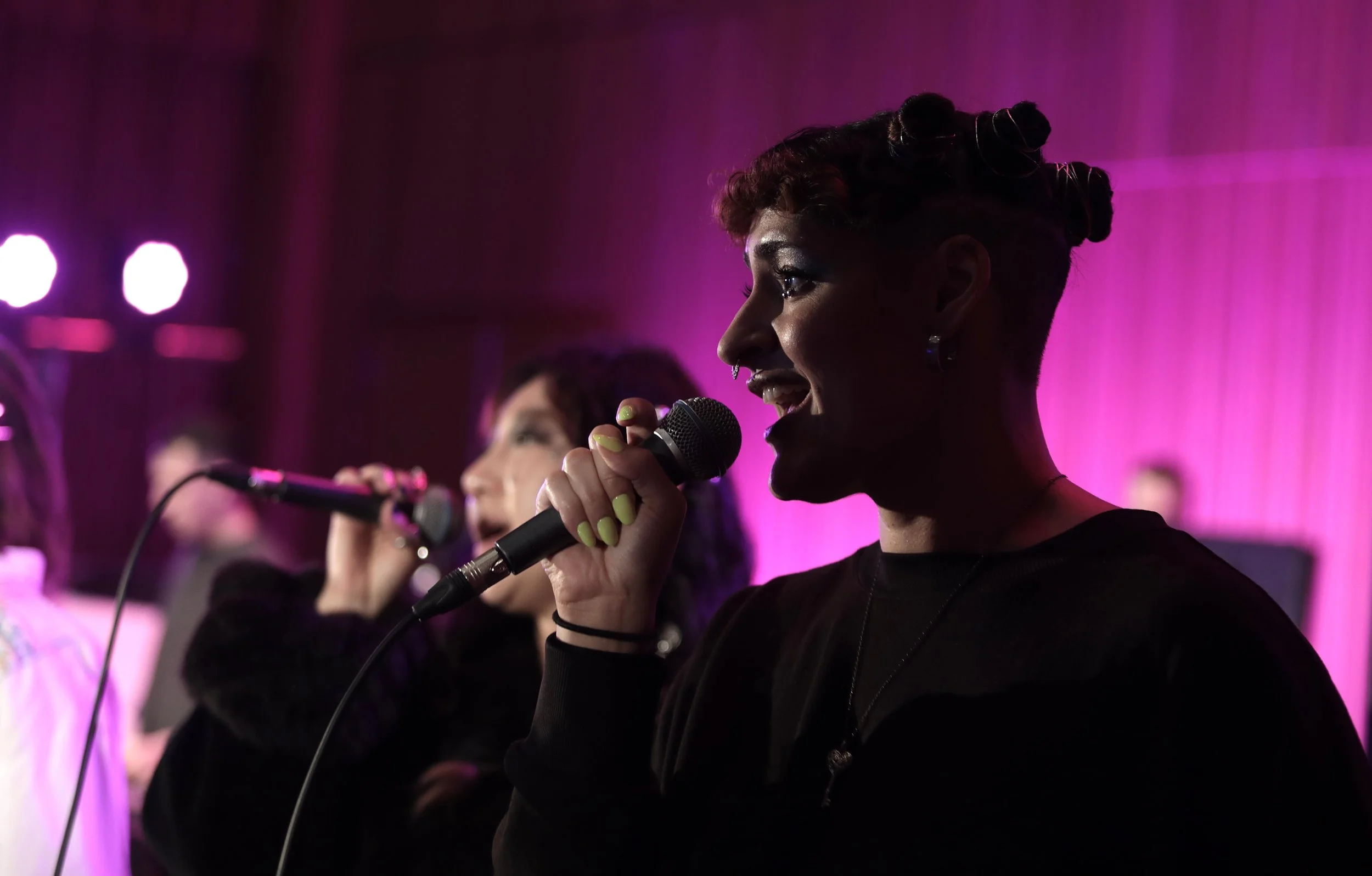 A woman with short hair and earrings singing into a microphone during a performance with pink and purple lighting.