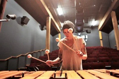 A woman wearing a beanie hat  playing a marimba with mallets in a theater.
