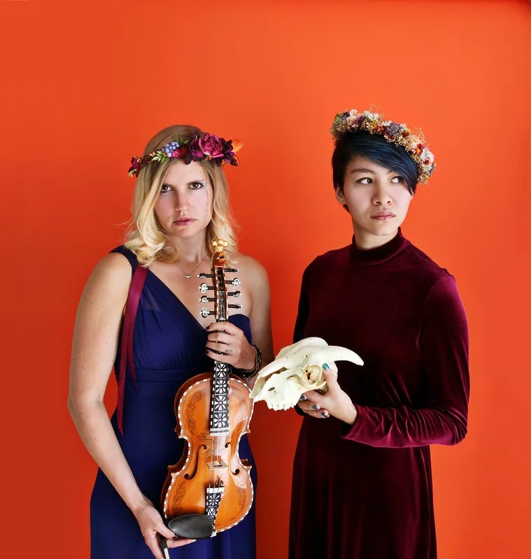 Two women wearing flower crowns against an orange background, one holding a violin and the other holding a skull.