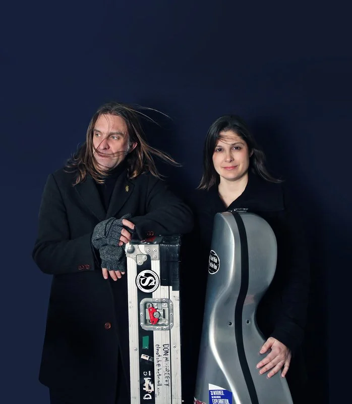 Two people, a man and a woman, with long hair, standing against a dark background, one with a guitar case and one with a cello case, both dressed in dark coats.