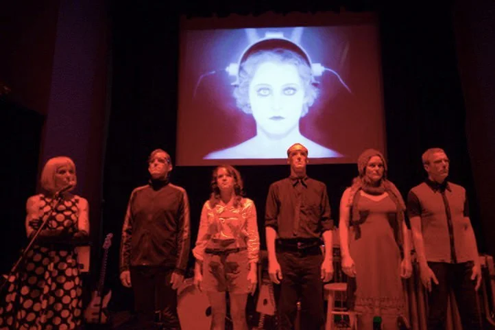 Six performers stand on stage in front of a projection of a woman's face with lightning effects, during a theatrical performance.