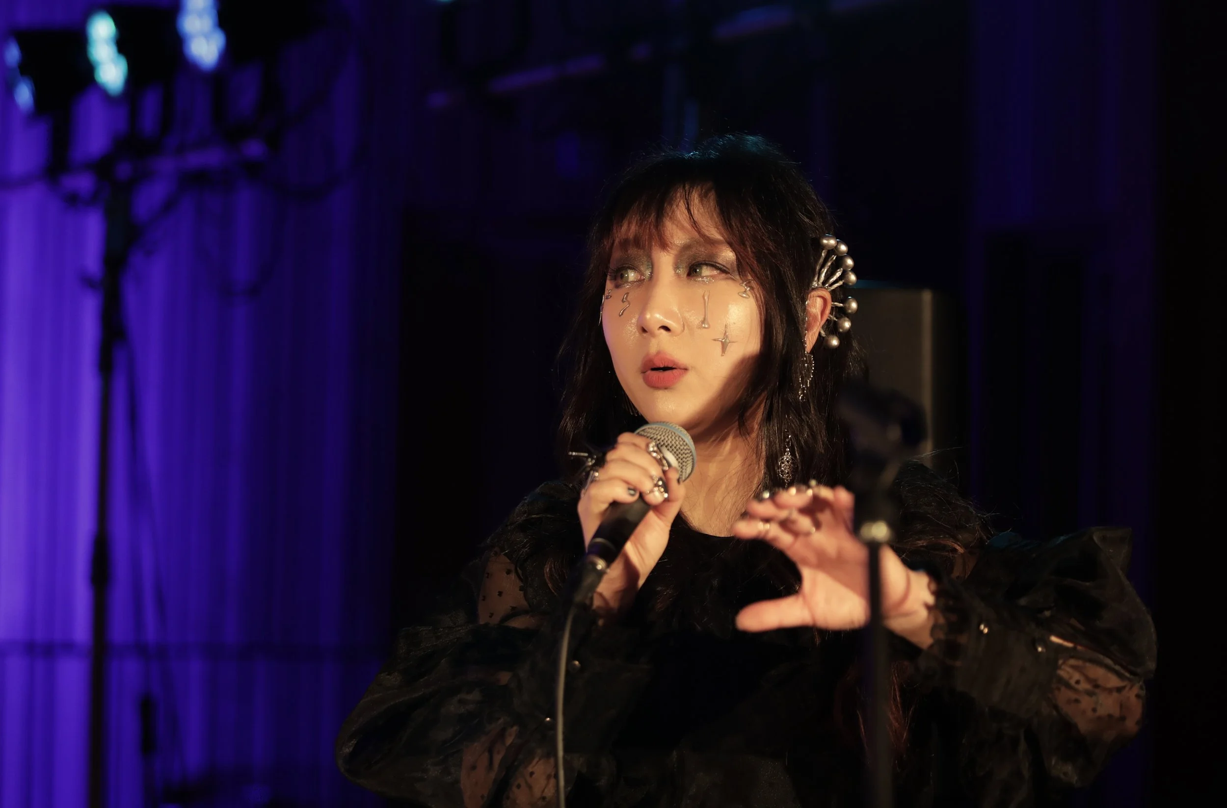 A woman on stage with dark hair and makeup, wearing a black lace outfit, holding a microphone, against a purple curtain background with stage lighting.