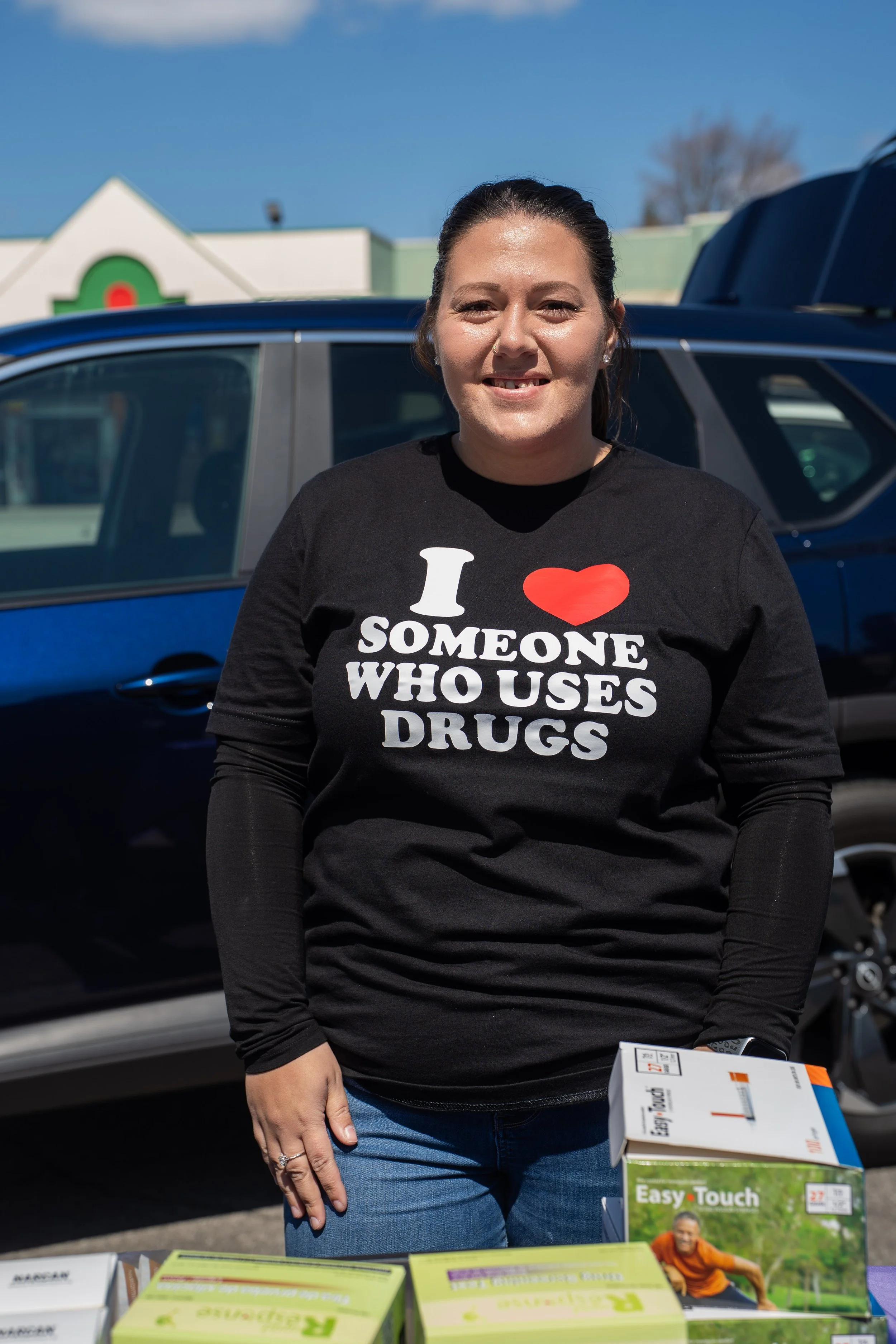 A woman standing outdoors in front of cars, wearing a black shirt with white and red text that says 'I ❤️ SOMEONE WHO USES DRUGS'. She is smiling and holding a box of Easy Touch diabetes test strips. There are banners and posters on a table in front of her.