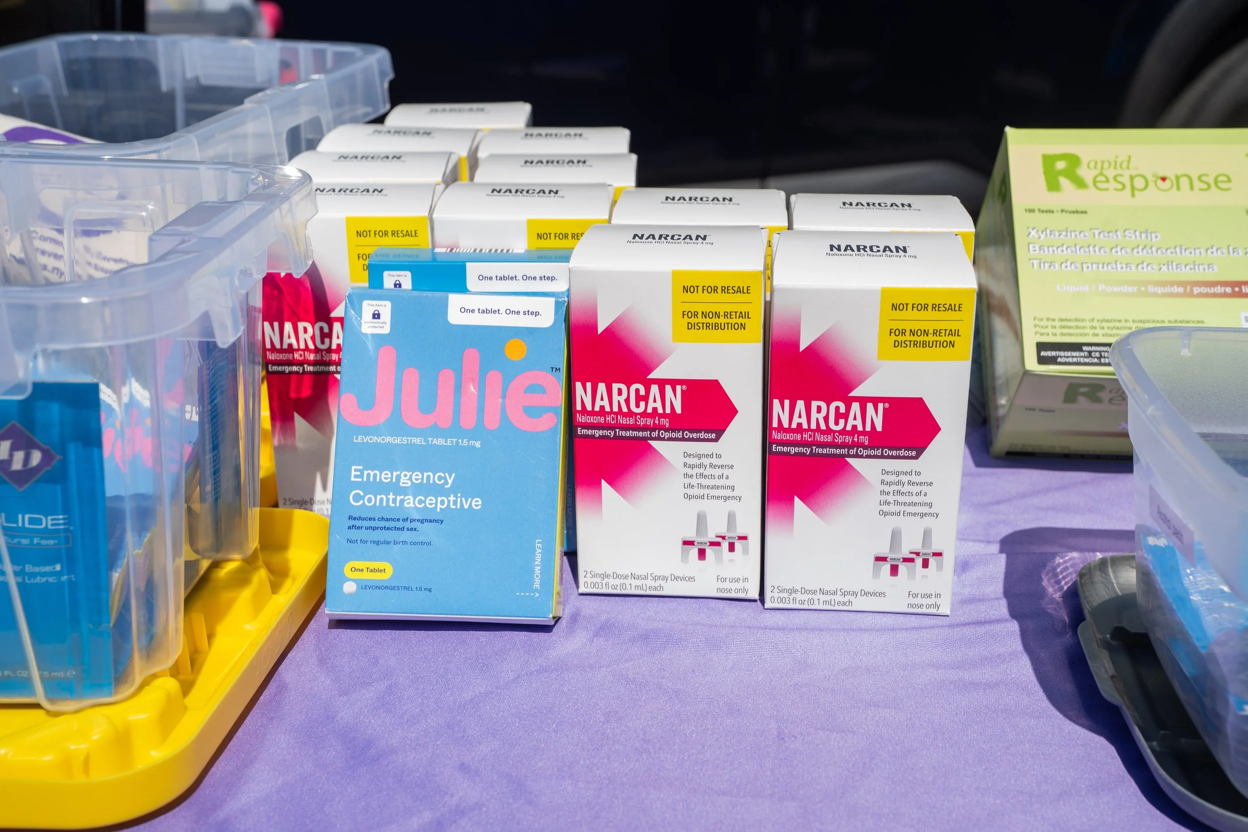 Display of emergency healthcare products including Narcan nasal spray boxes, Julie contraceptive tablets, and Rapid Response test strips on a purple tablecloth.