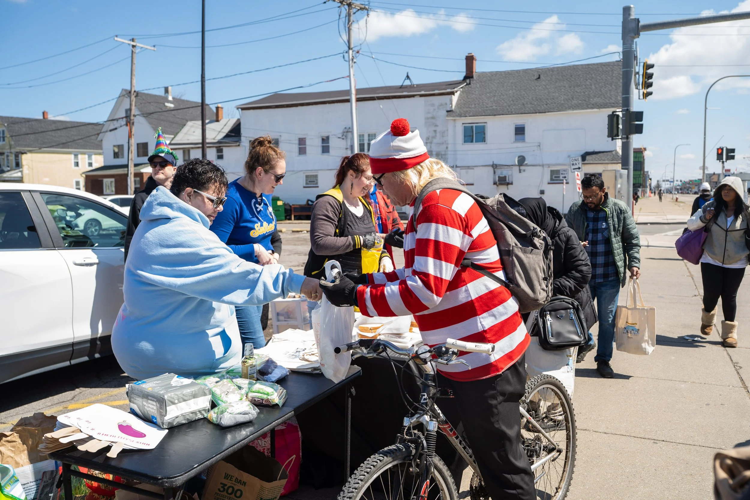 A group of people gathered around a table outdoors on a city sidewalk, with some wearing masks and casual clothing, including a man in a red and white striped shirt and Santa hat, and another person with a party hat, possibly participating in a community event or donation drive.