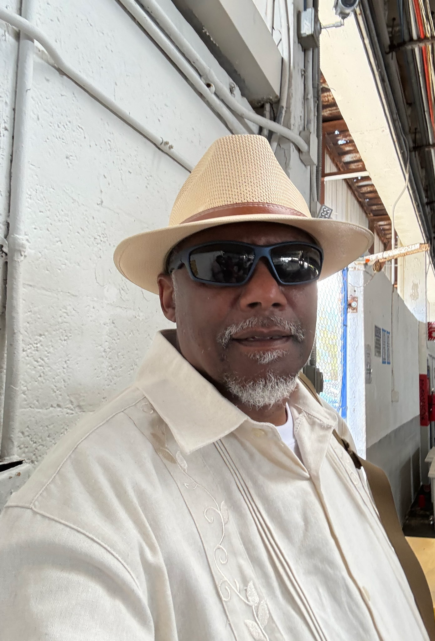 A man wearing a beige hat, dark sunglasses, and a beige embroidered shirt, standing indoors against a white wall with visible pipes and wiring.