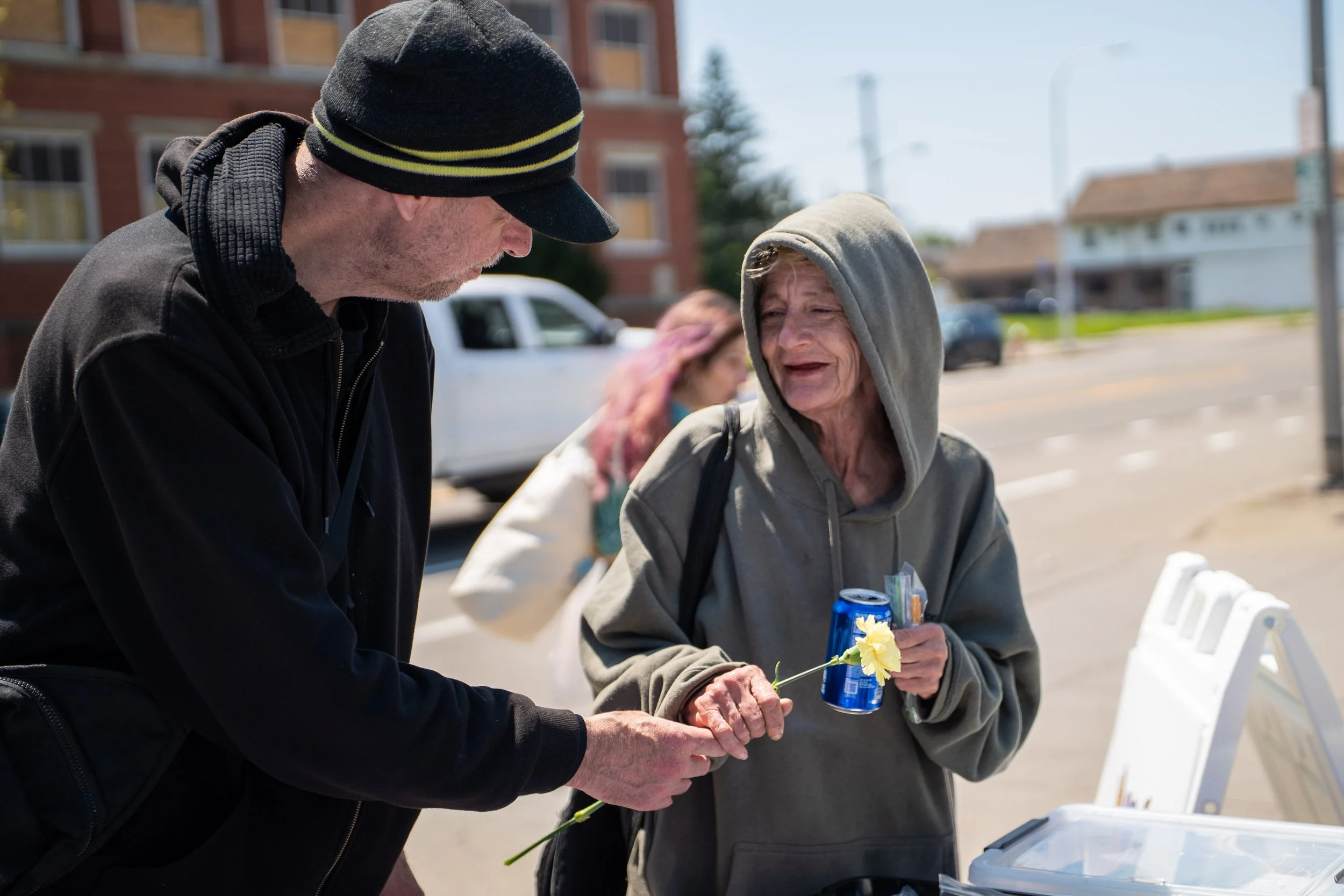 A man with a black cap and black hoodie holding a yellow flower and talking to an elderly woman in a gray hoodie who is holding a soda can and a yellow flower.