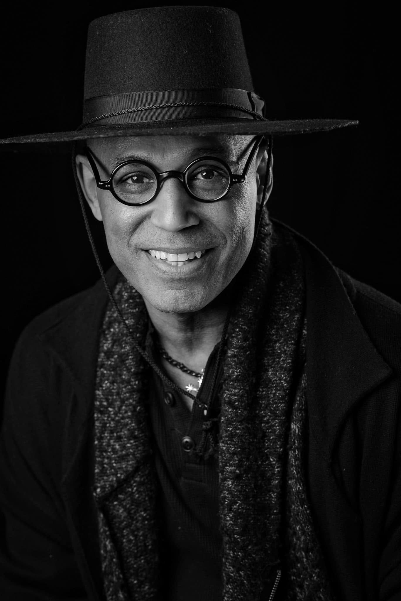 Black and white portrait of a man with glasses wearing a wide-brimmed hat and a patterned scarf, smiling slightly.